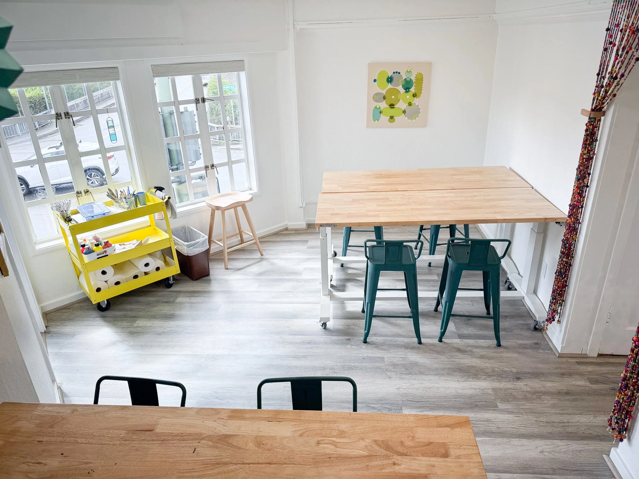 An art classroom with large windows, a yellow rolling cart with art supplies, a small wooden stool, and a large wooden table with teal chairs. A colorful beaded curtain is on the right and a whimsical artwork on the wall.