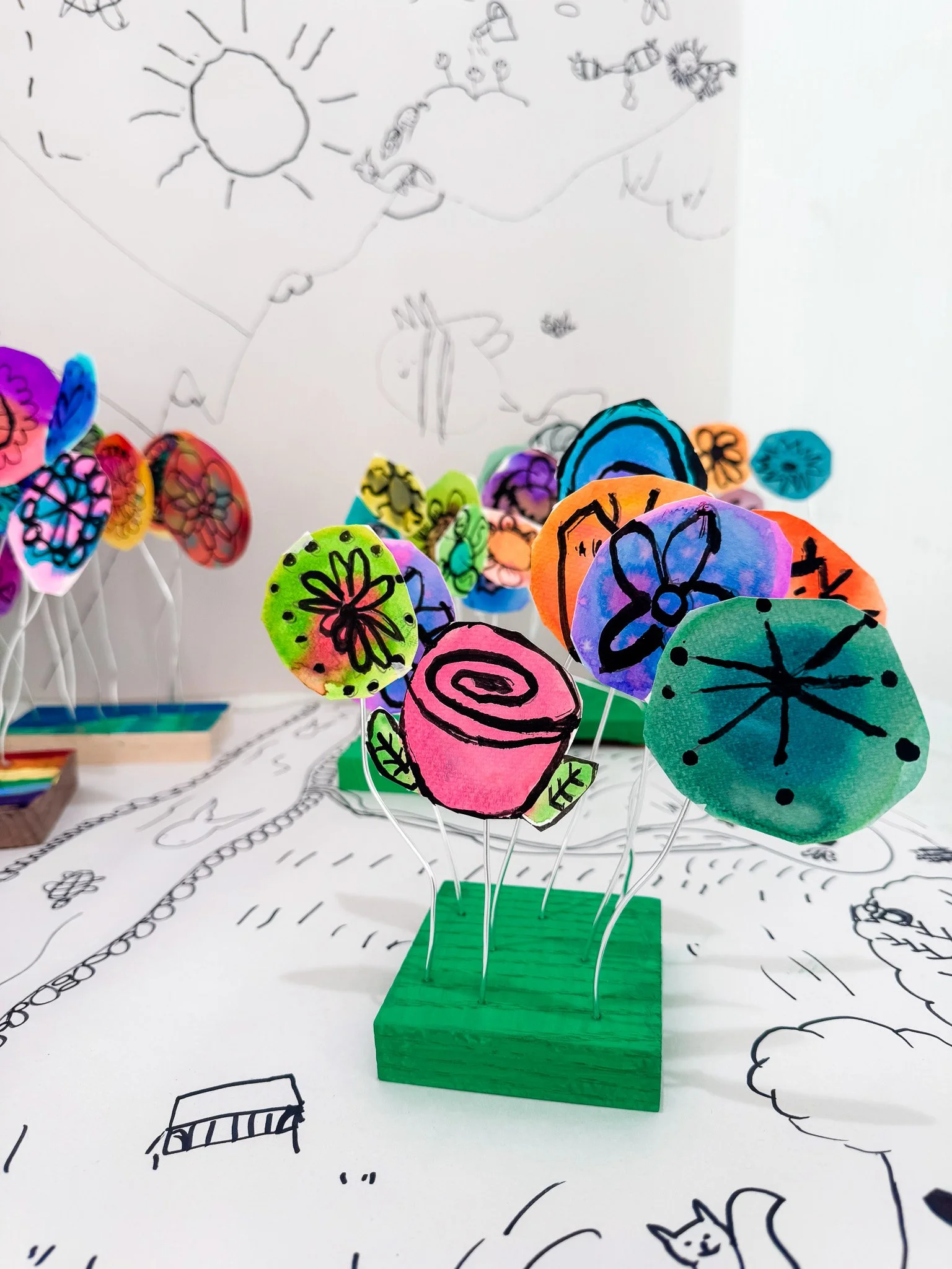 Colorful paper flowers with black marker designs on the petals, arranged on thin wires in a green wooden base, with black and white drawings on the background
