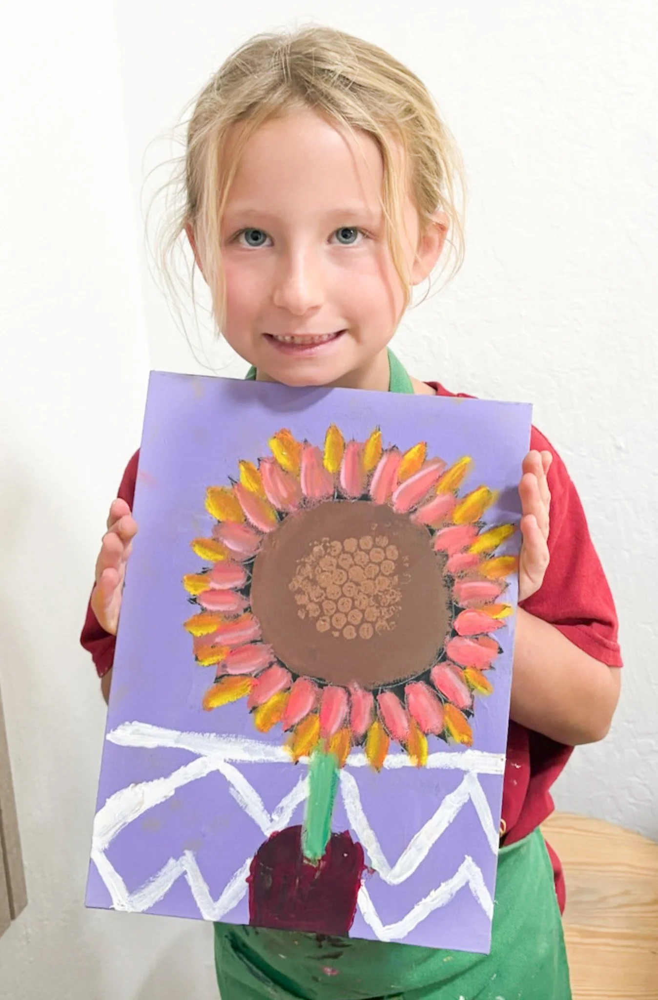 A young girl with blonde hair and a red shirt holds up her painted artwork of a sunflower on purple paper.