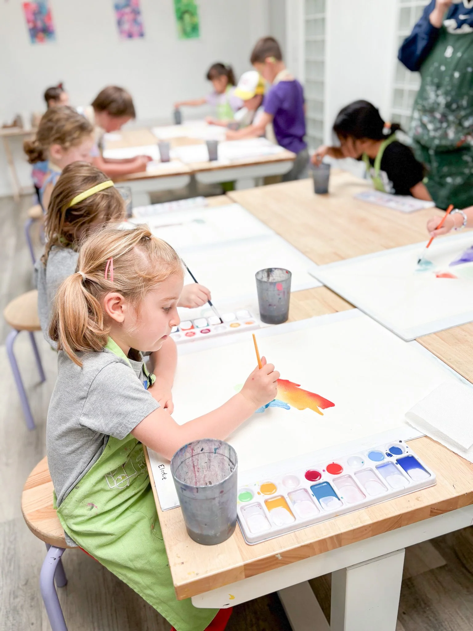 Children sitting at a table painting watercolor art in an art classroom.
