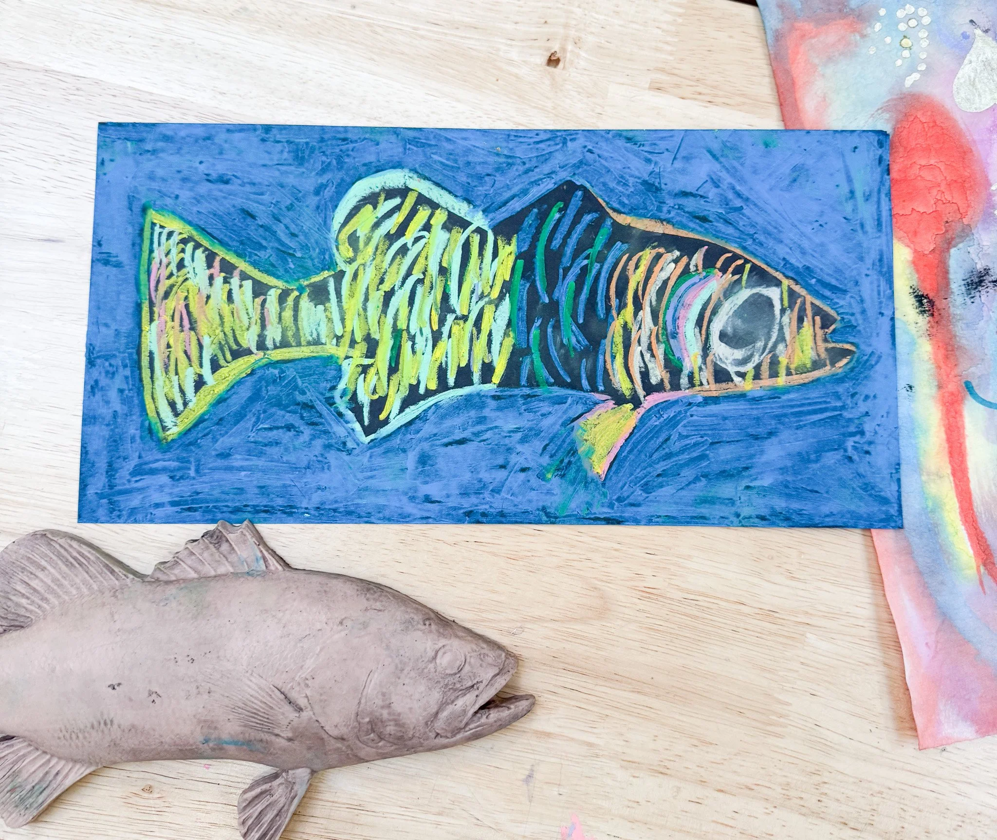 Colorful painting of a fish with yellow, green, and black lines on a blue background, alongside a real fish with grayish-white coloring on a wooden surface, partially visible.