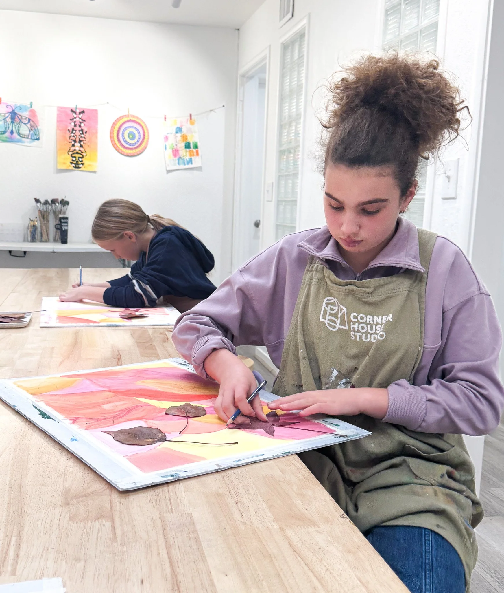 Two young girls sitting at a table working on art projects, with colorful artwork hanging on the wall in the background.