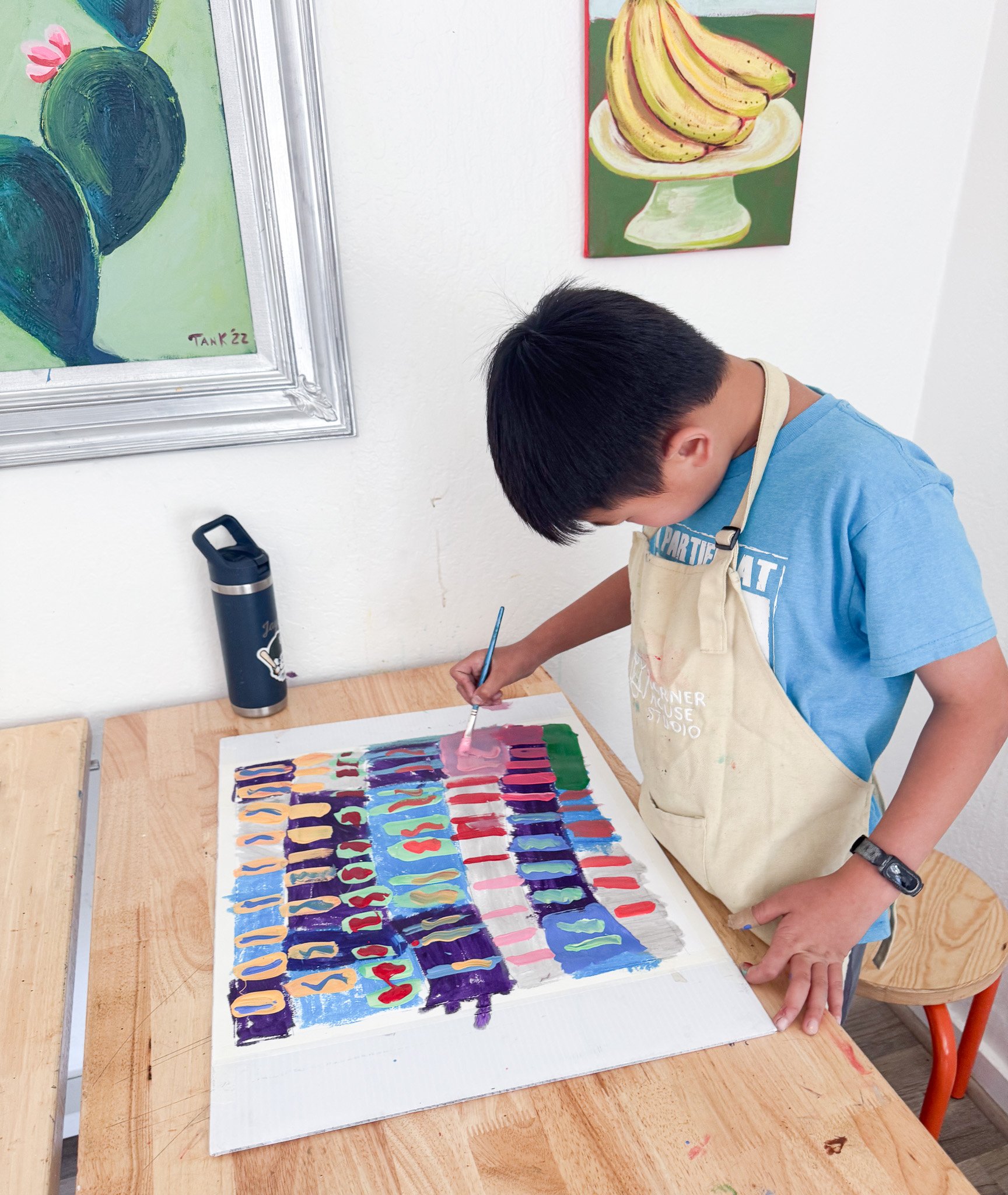 A boy painting on a large canvas with colorful patterns in an art studio, with paintings of cactus and bananas on the wall behind him.