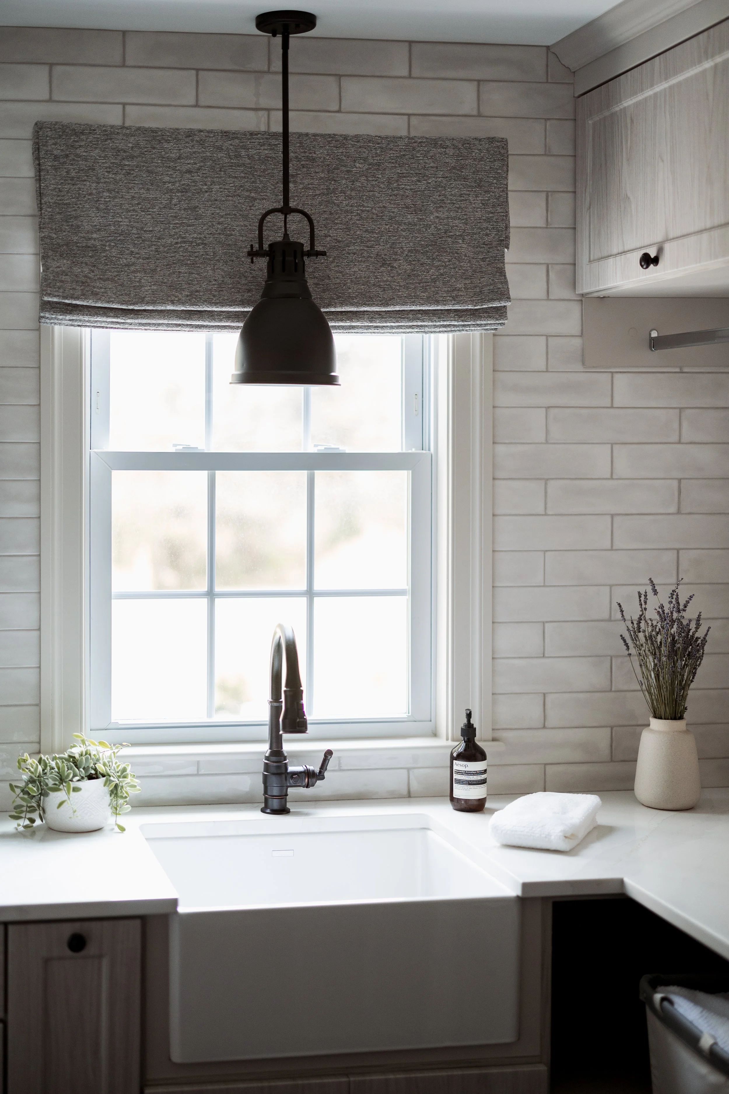 Modern kitchen sink with white basin, black faucet, and large window. Gray Roman shade covers the top of the window. A black pendant light hangs above. On the countertop, there’s a bottle of soap, a white towel, potted plant, and vase with lavender. 