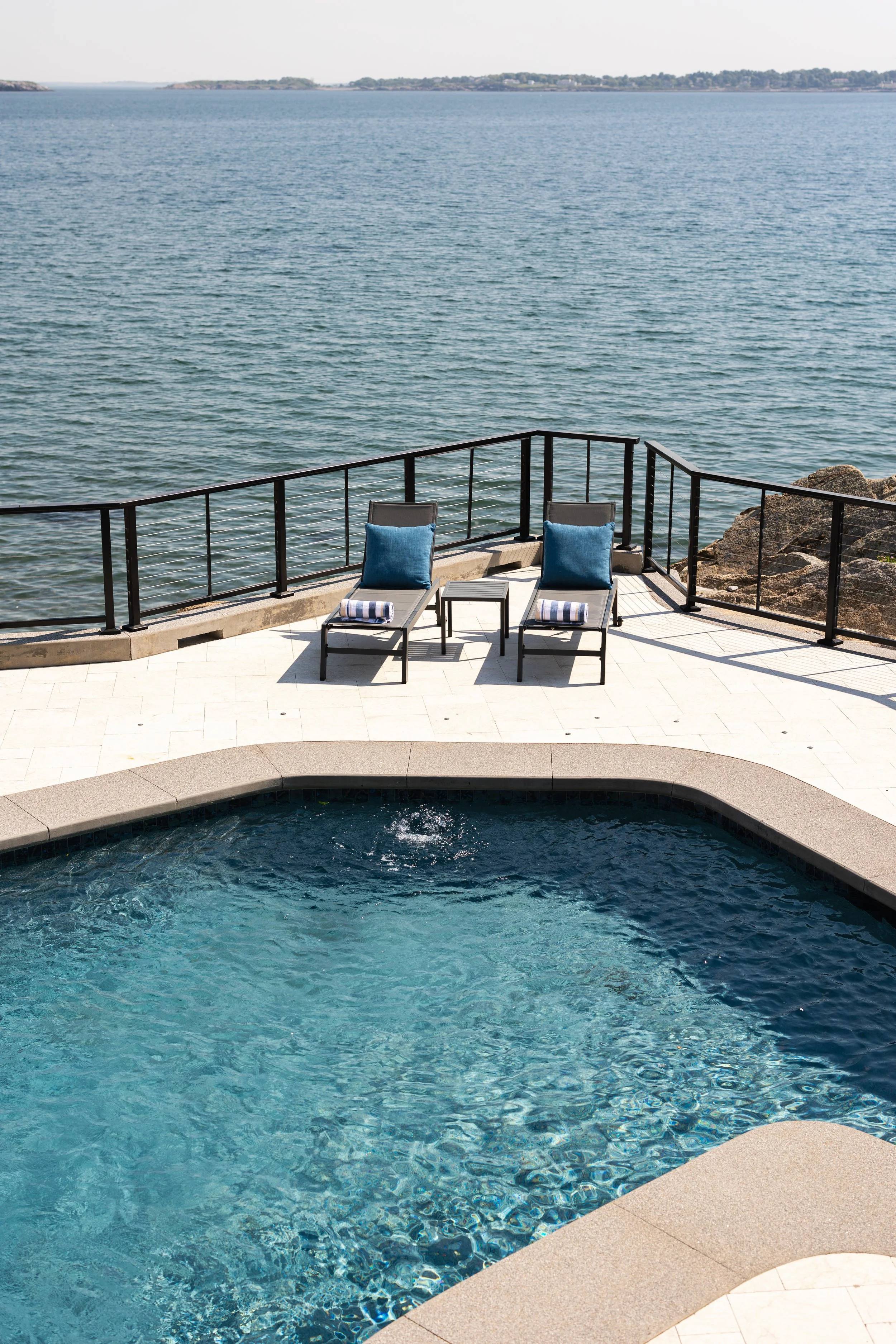 View of a swimming pool with two lounge chairs and a table overlooking a body of water, enclosed by a black metal railing.
