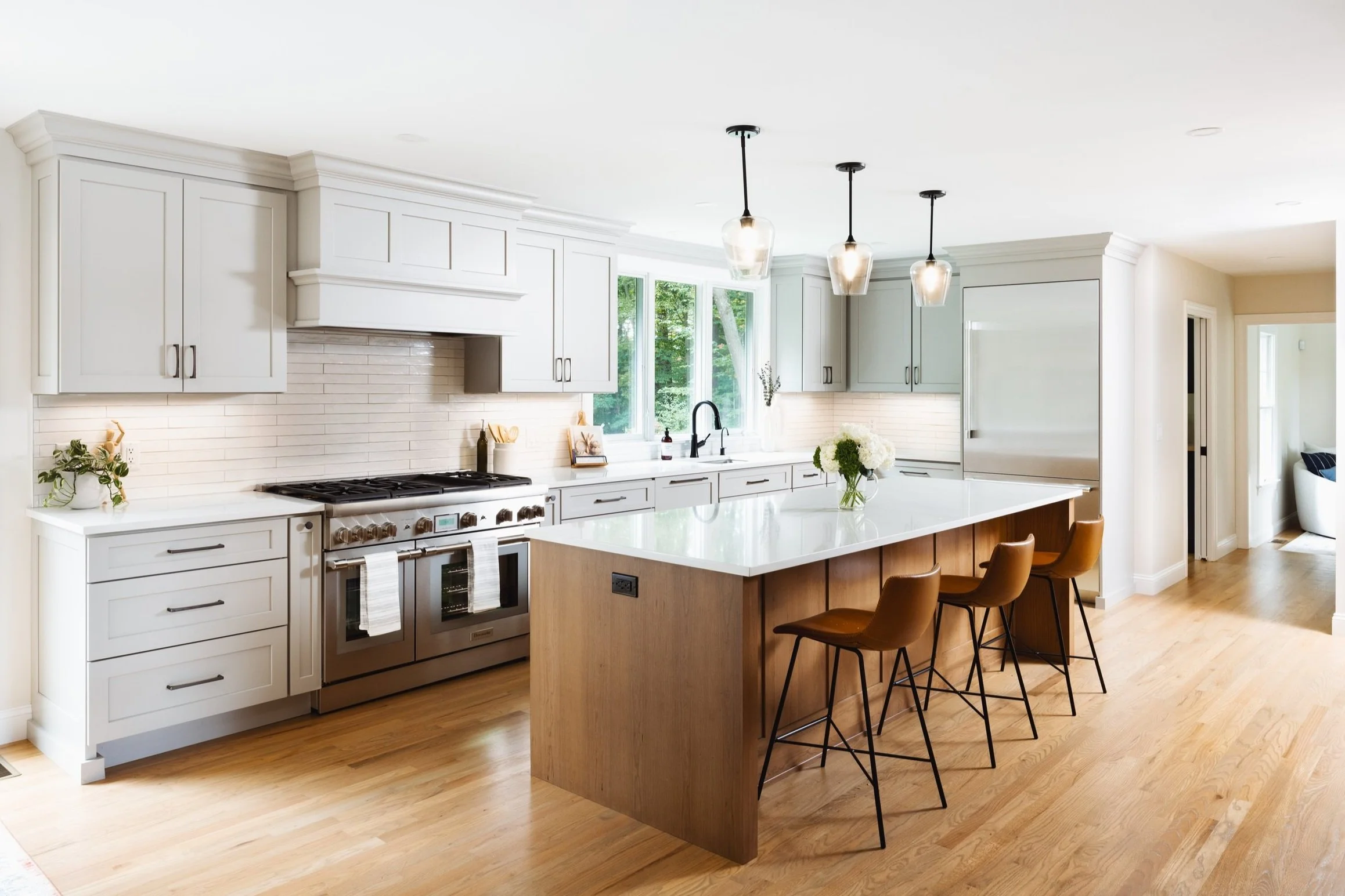 Modern kitchen with white cabinets, a large center island with wooden base, marble countertop, and brown chairs. Stainless steel appliances, pendant lights, and hardwood floors.