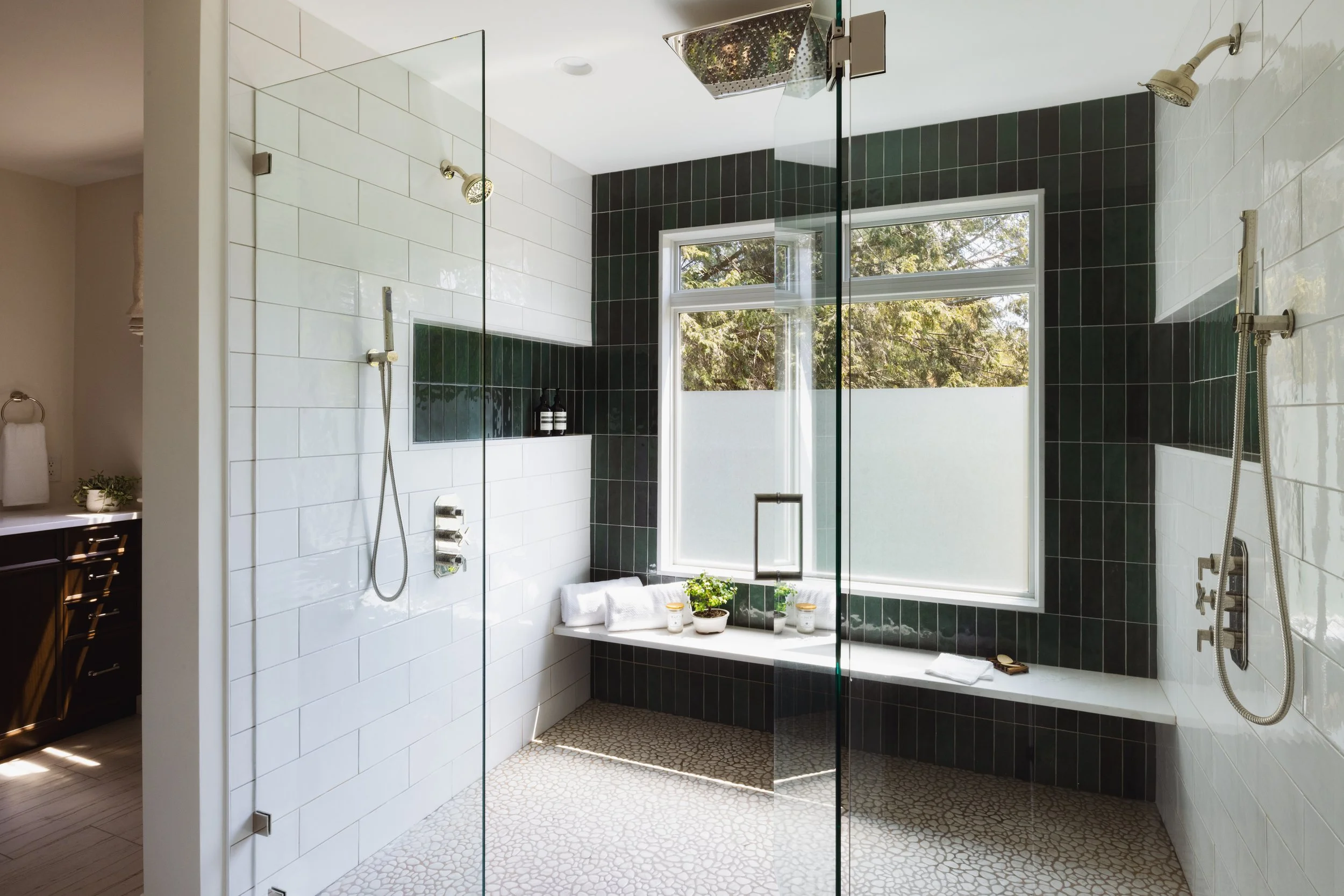 Modern bathroom with glass shower, white and dark green tiles, rainfall showerhead, and bench with plants and towels; window with frosted glass.