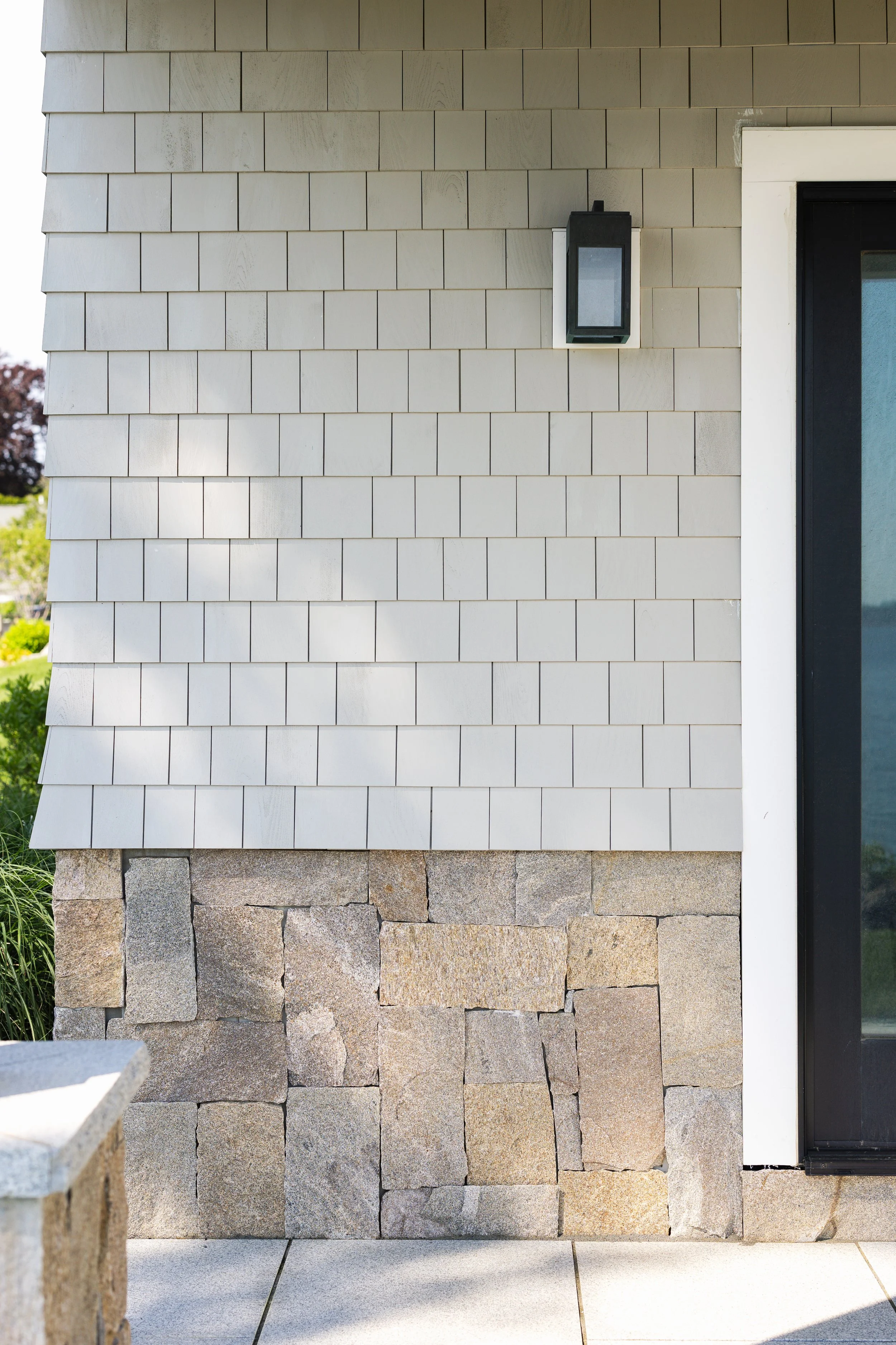 Exterior house wall with shingles and stone, featuring a black outdoor light fixture next to a black-framed window.