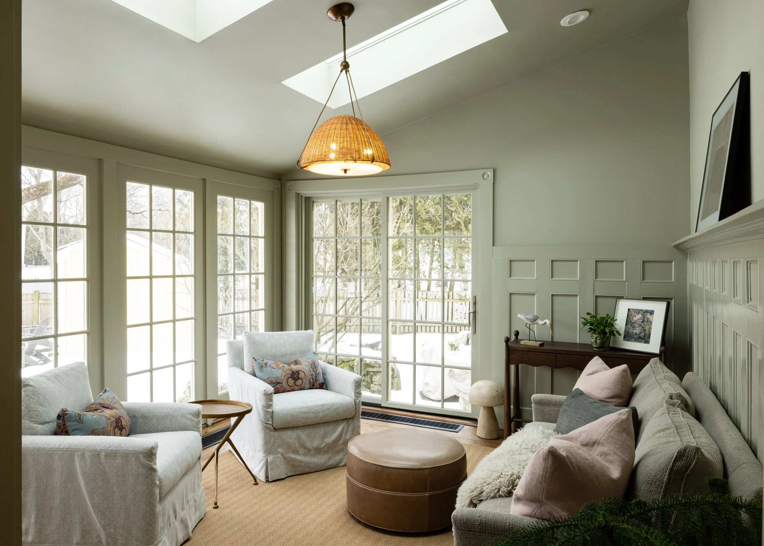 Cozy sunroom with large windows, two armchairs, a sofa, round ottoman, small side table, and pendant light. Wall panels and a sliding glass door lead to a snowy outdoor area.