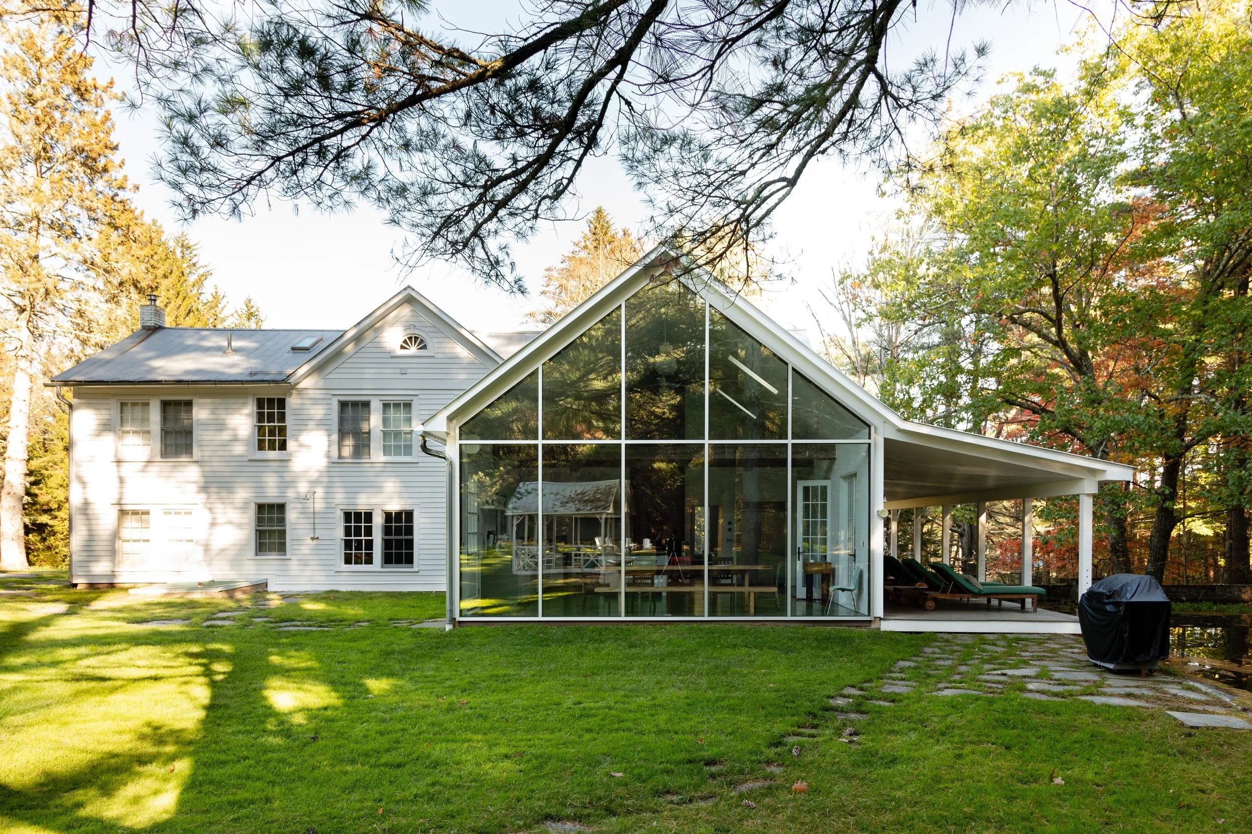 Modern house with large glass extension in a wooded setting; original structure is white and traditional; green lawn and outdoor furniture visible.