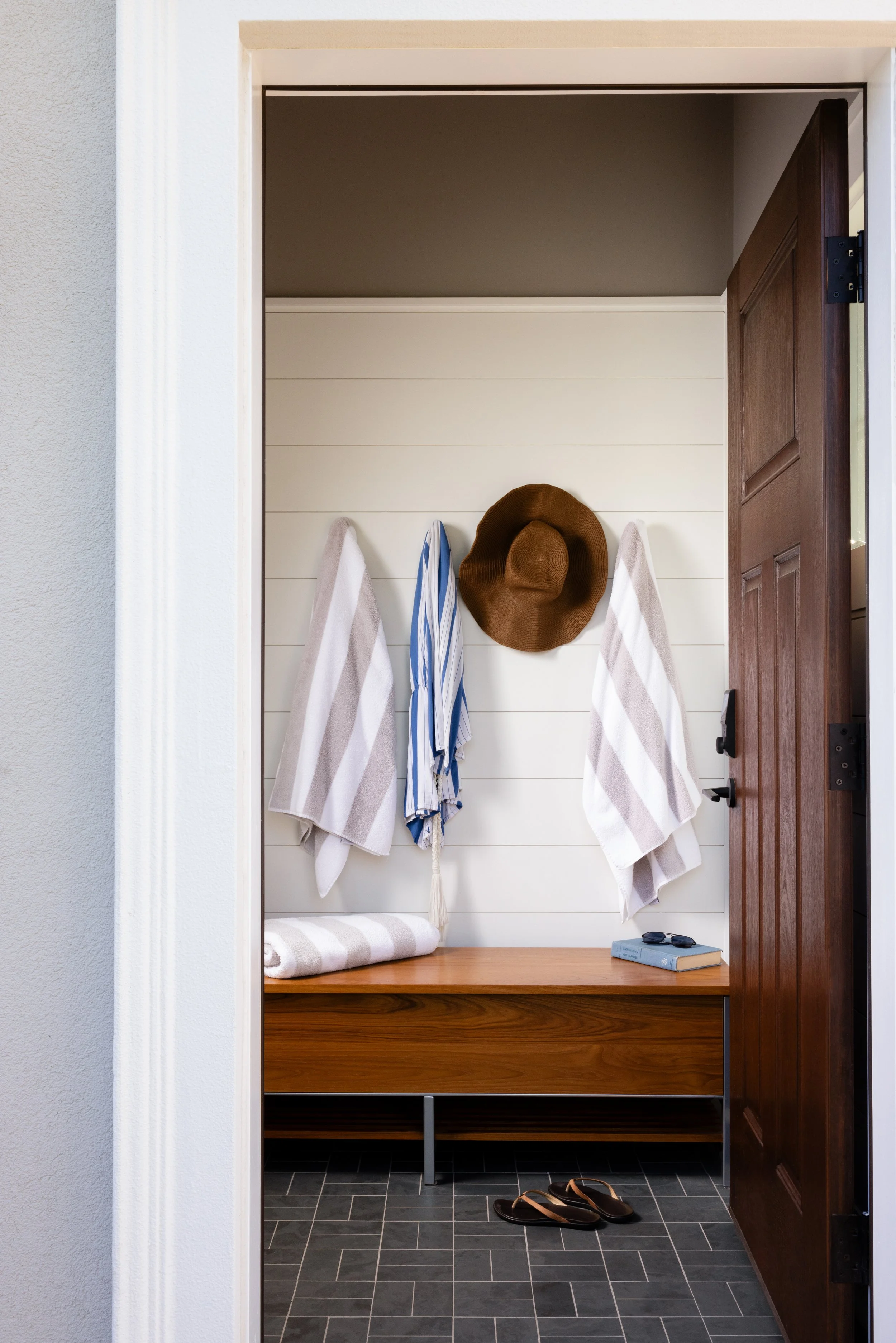 Entrance with wooden bench, striped towels, straw hat on hooks, slippers on tiled floor, and open door.