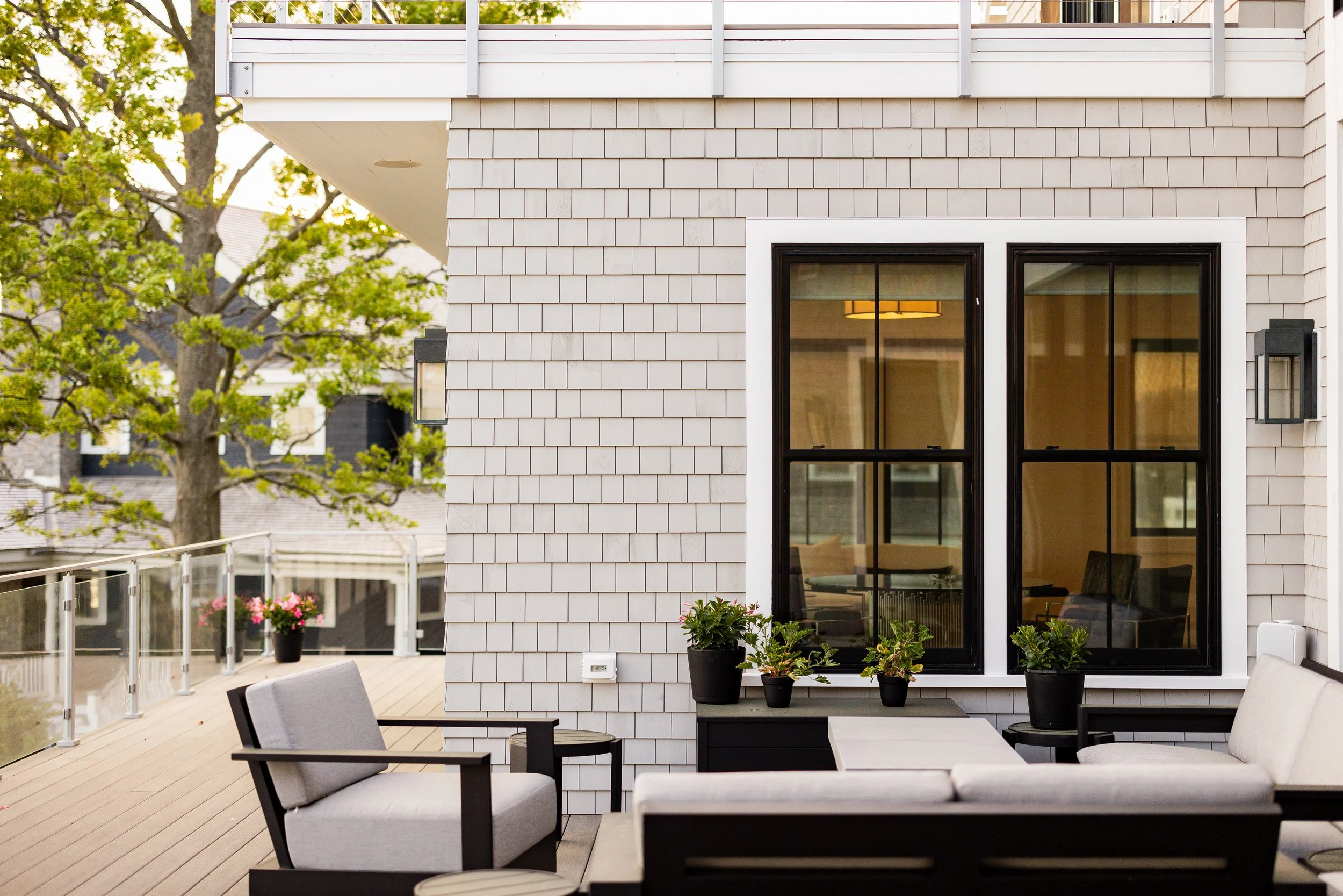 Modern outdoor patio with gray wicker furniture and potted plants on a wooden deck, adjacent to a house with shingle siding and large windows, surrounded by trees.
