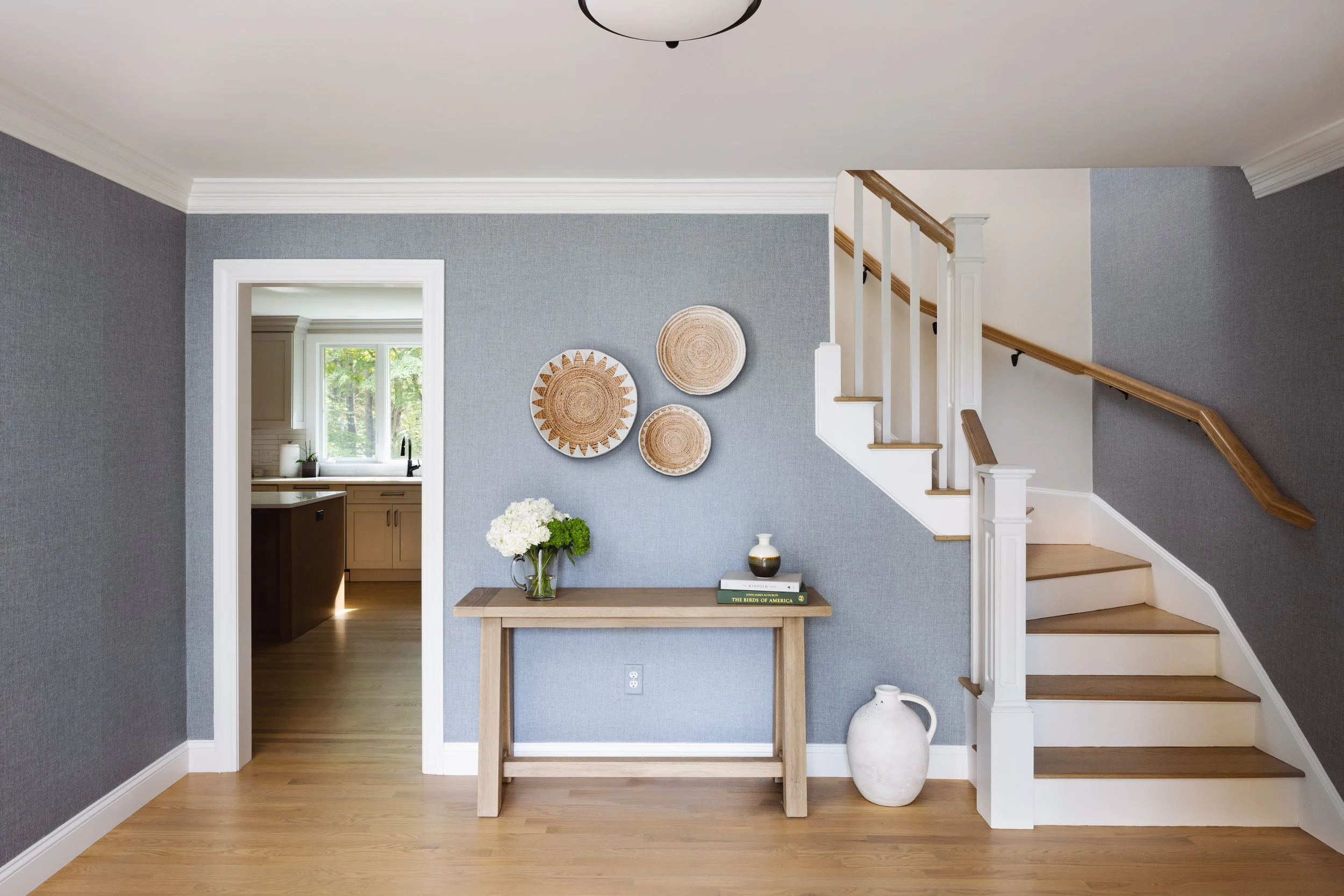 Modern foyer with wooden stairs, blue textured walls, a console table, decorative baskets on the wall, and a view into a kitchen.