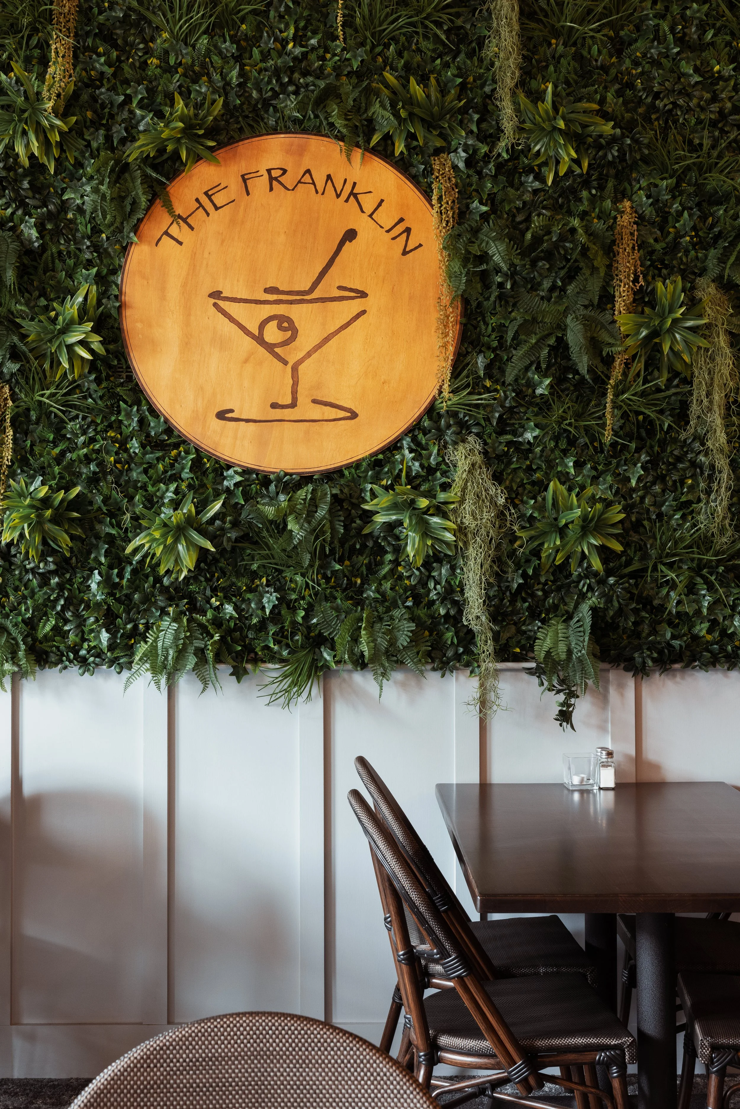Interior of a restaurant with a leafy green wall and a circular wooden sign displaying a martini glass illustration and the text 'The Franklin.' Below are dark wooden chairs and a table partially visible.