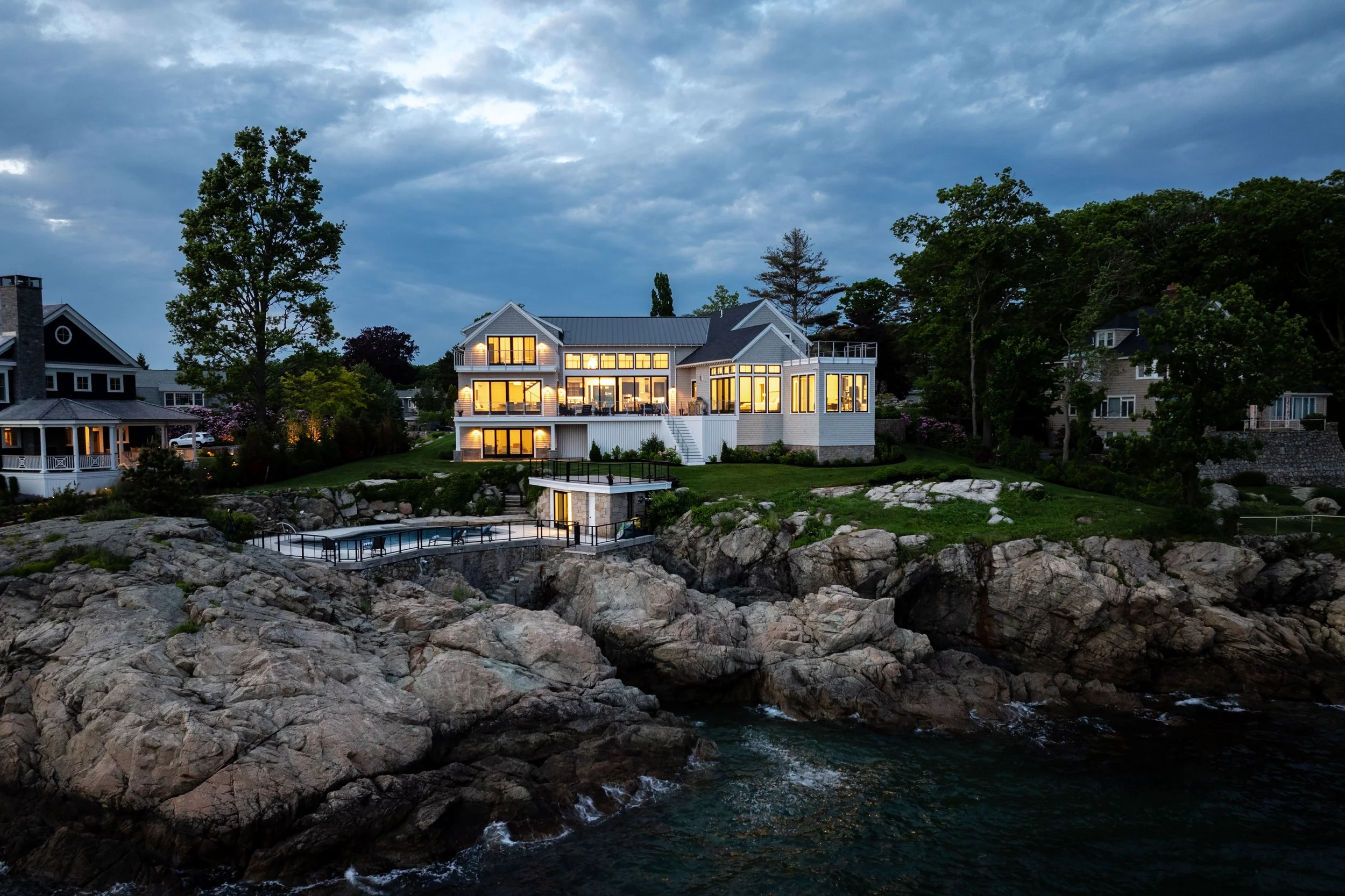 A large modern coastal home with illuminated windows, situated on rocky cliffs, overlooking the ocean at dusk.