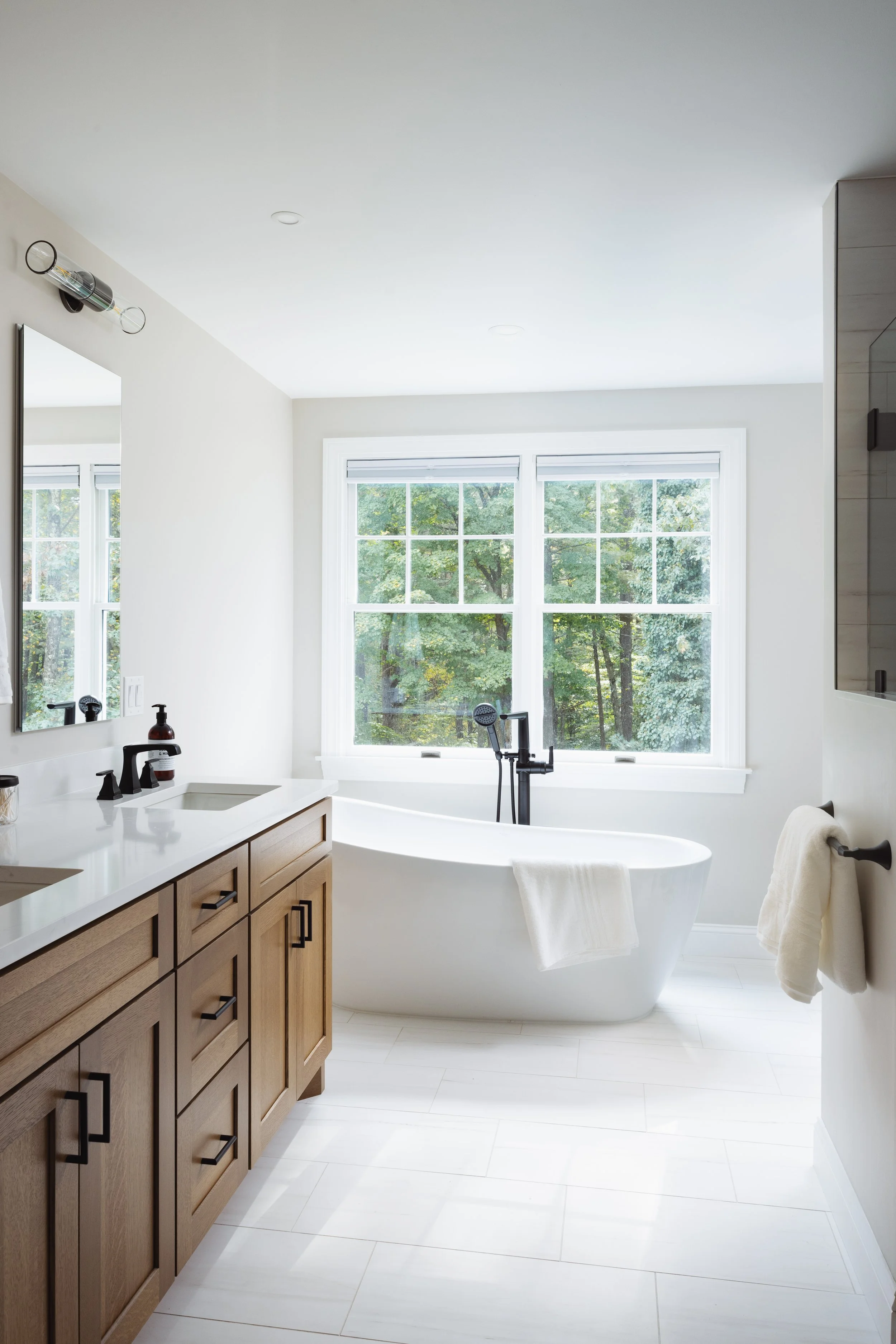 Modern bathroom with freestanding bathtub, wooden vanity, large window, and white tiled floor.