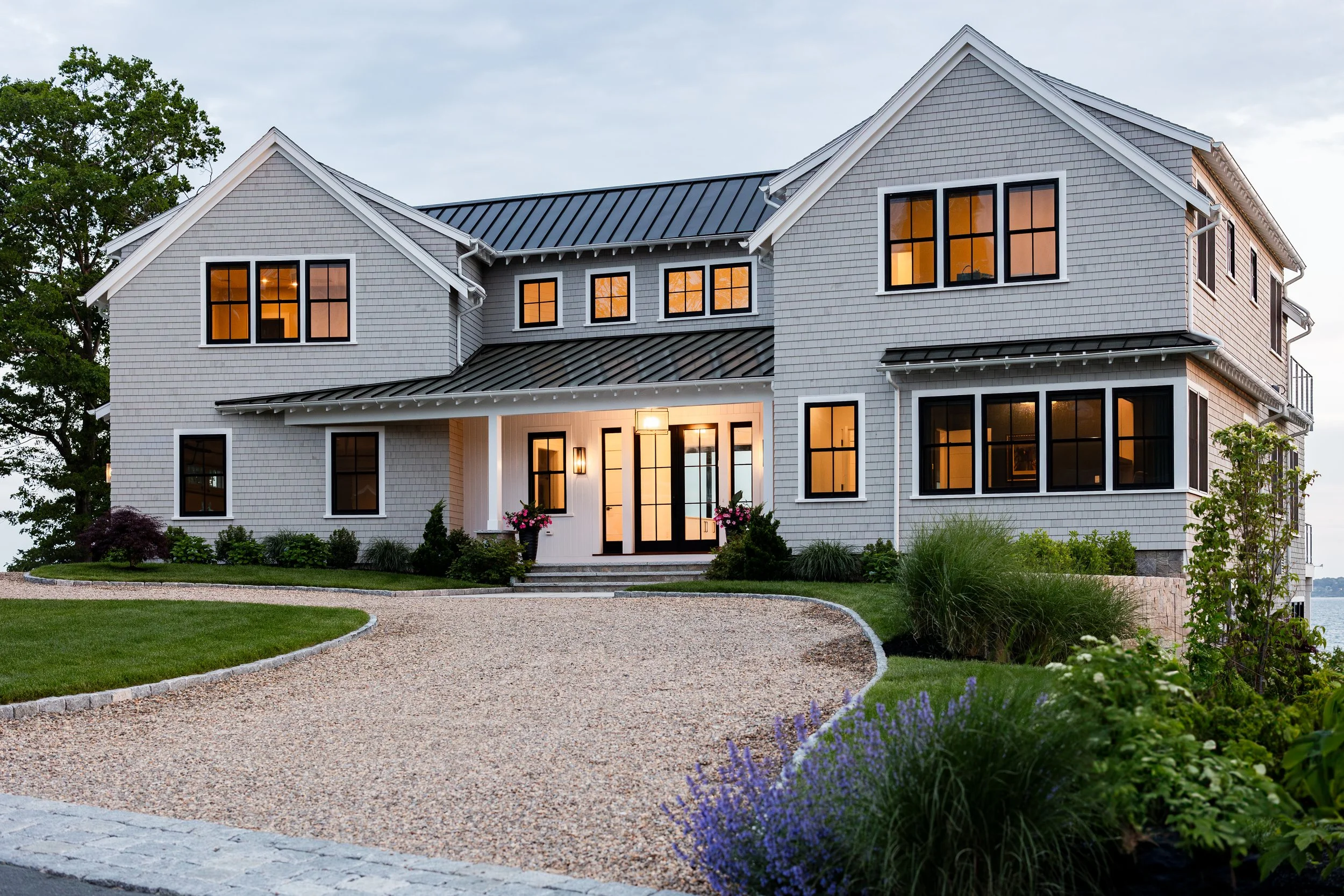 Modern two-story house with gray siding and a metal roof, featuring numerous illuminated windows, a gravel driveway, landscaped garden, and surrounding greenery.
