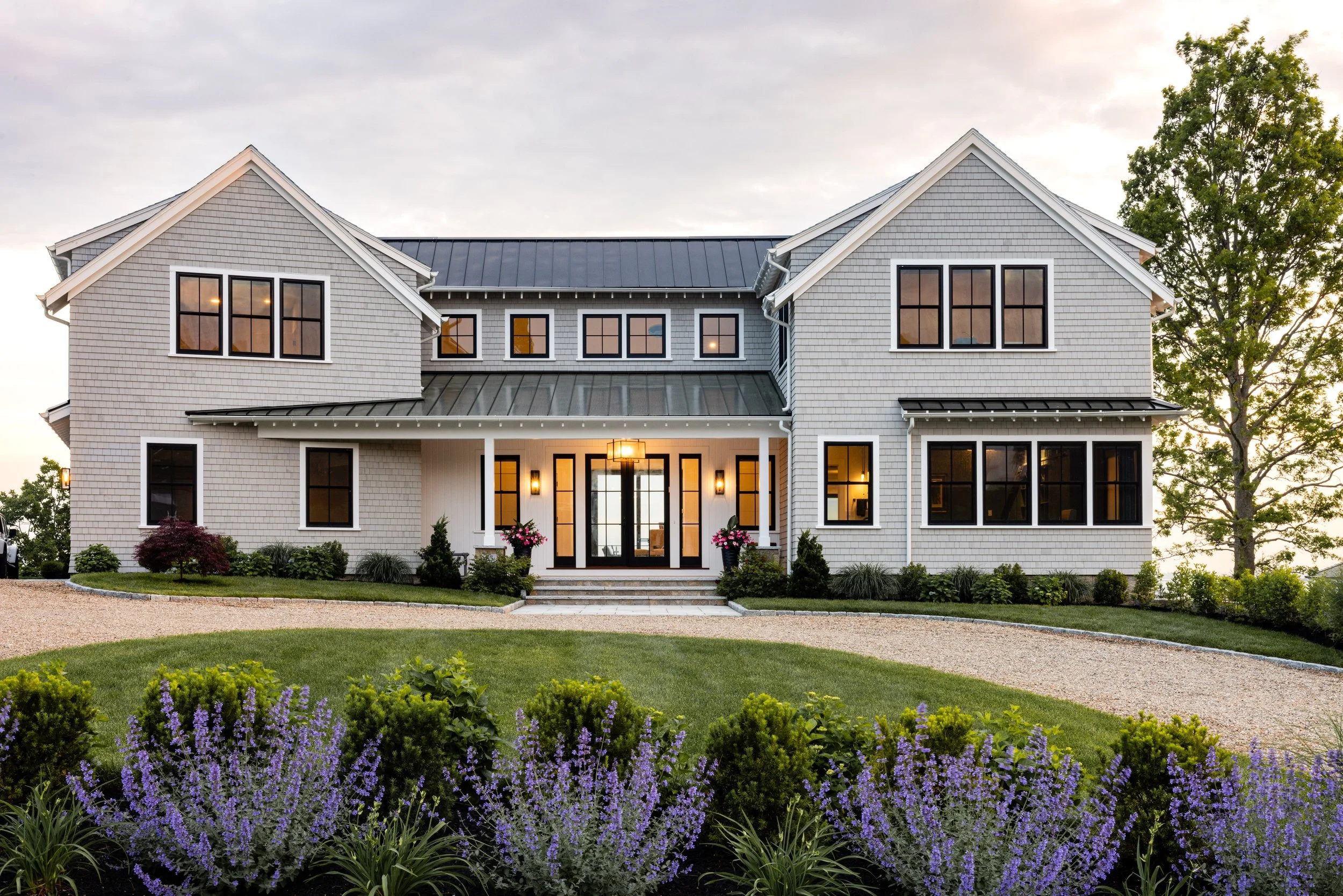 Modern two-story house with gray siding, black framed windows, metal roof, front porch, landscaped garden with purple flowers, and gravel driveway.
