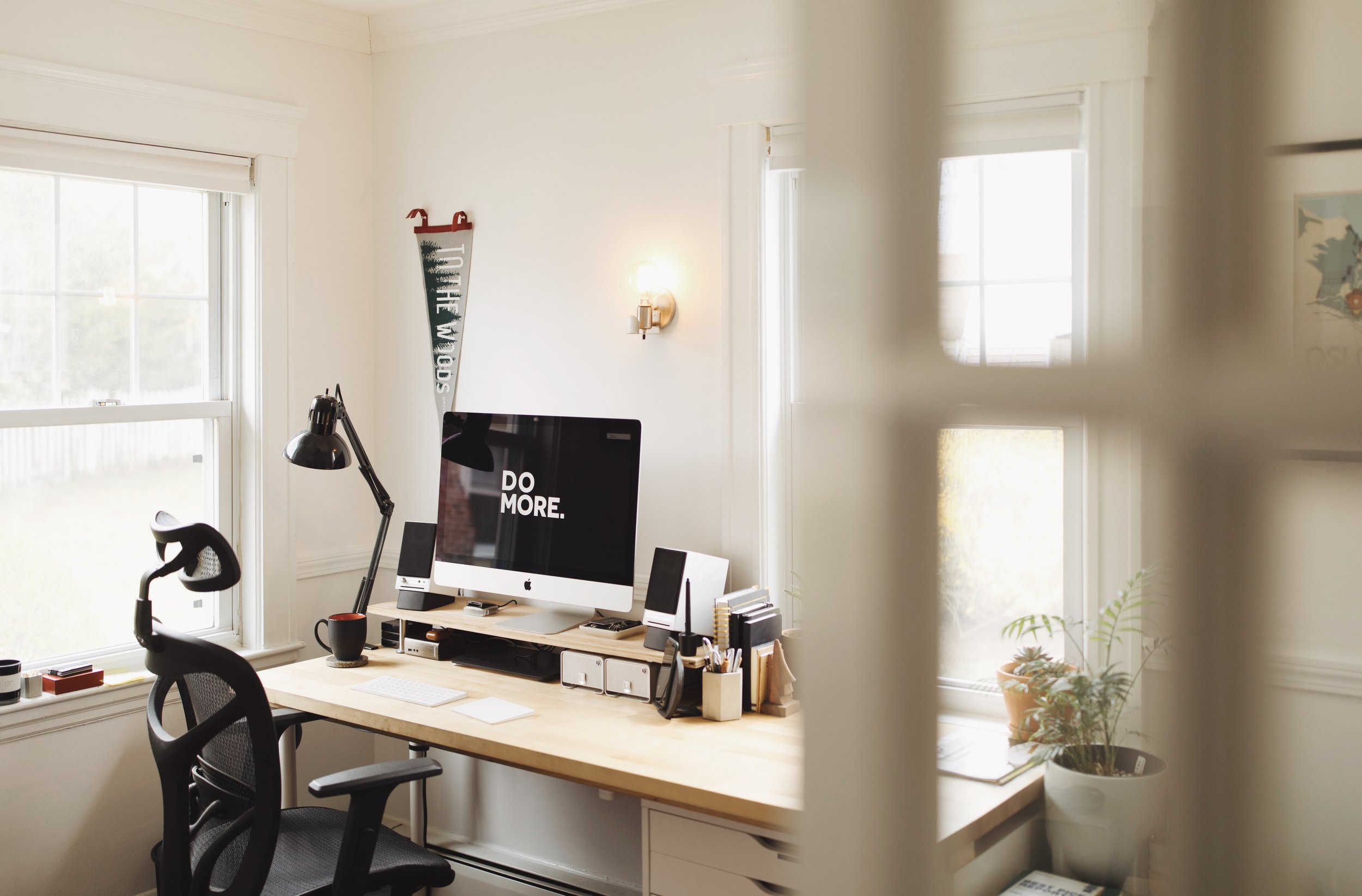 A home office with a wooden desk, computer monitor displaying 'Do More,' ergonomic chair, desk lamp, and office supplies. Light streams through two large windows. A potted plant and a decorative wall piece are also visible.