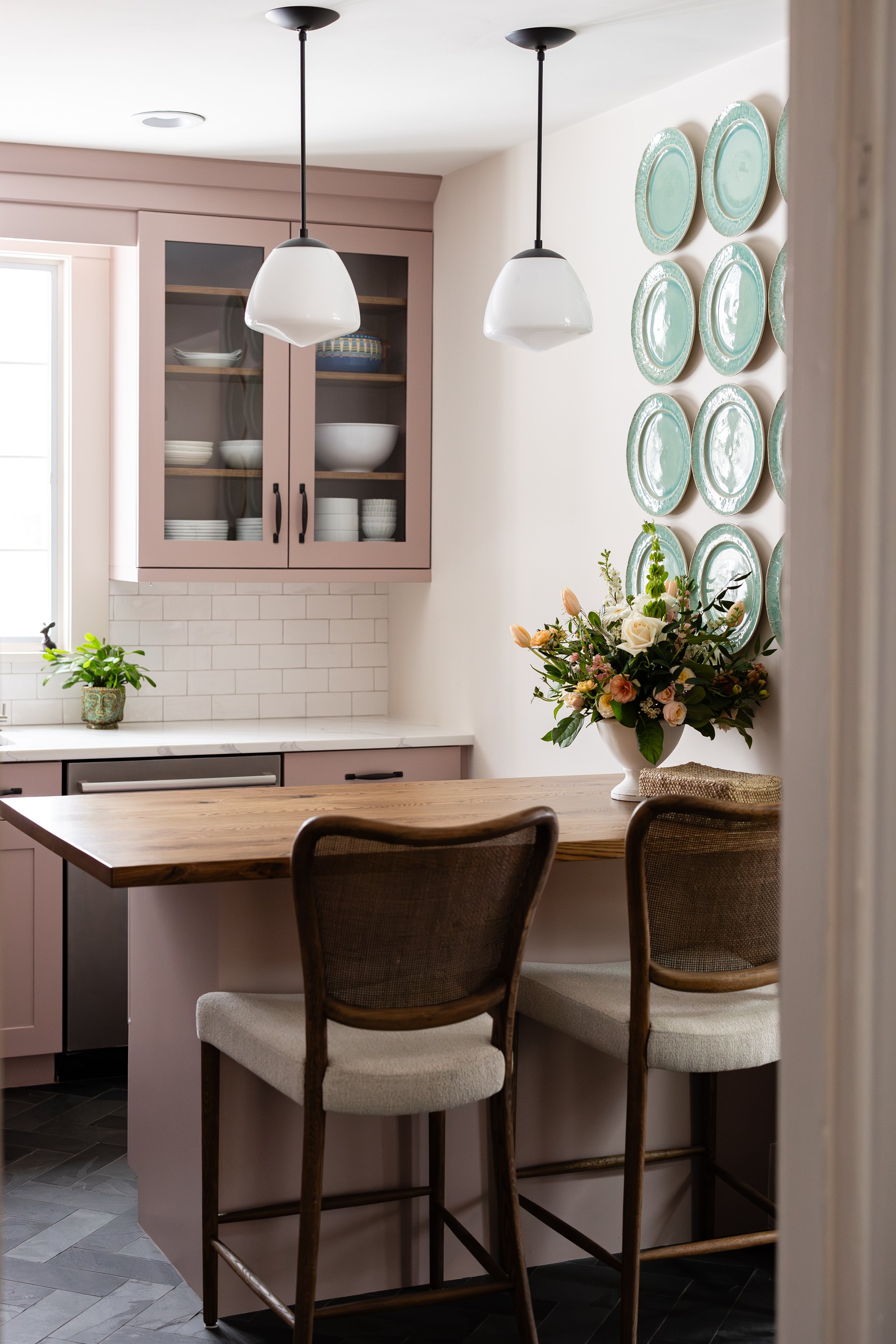 Cozy kitchen with barstools, wooden countertop, pendant lights, floral arrangement, decorative plates on wall, pink cabinets, and subway tile backsplash.
