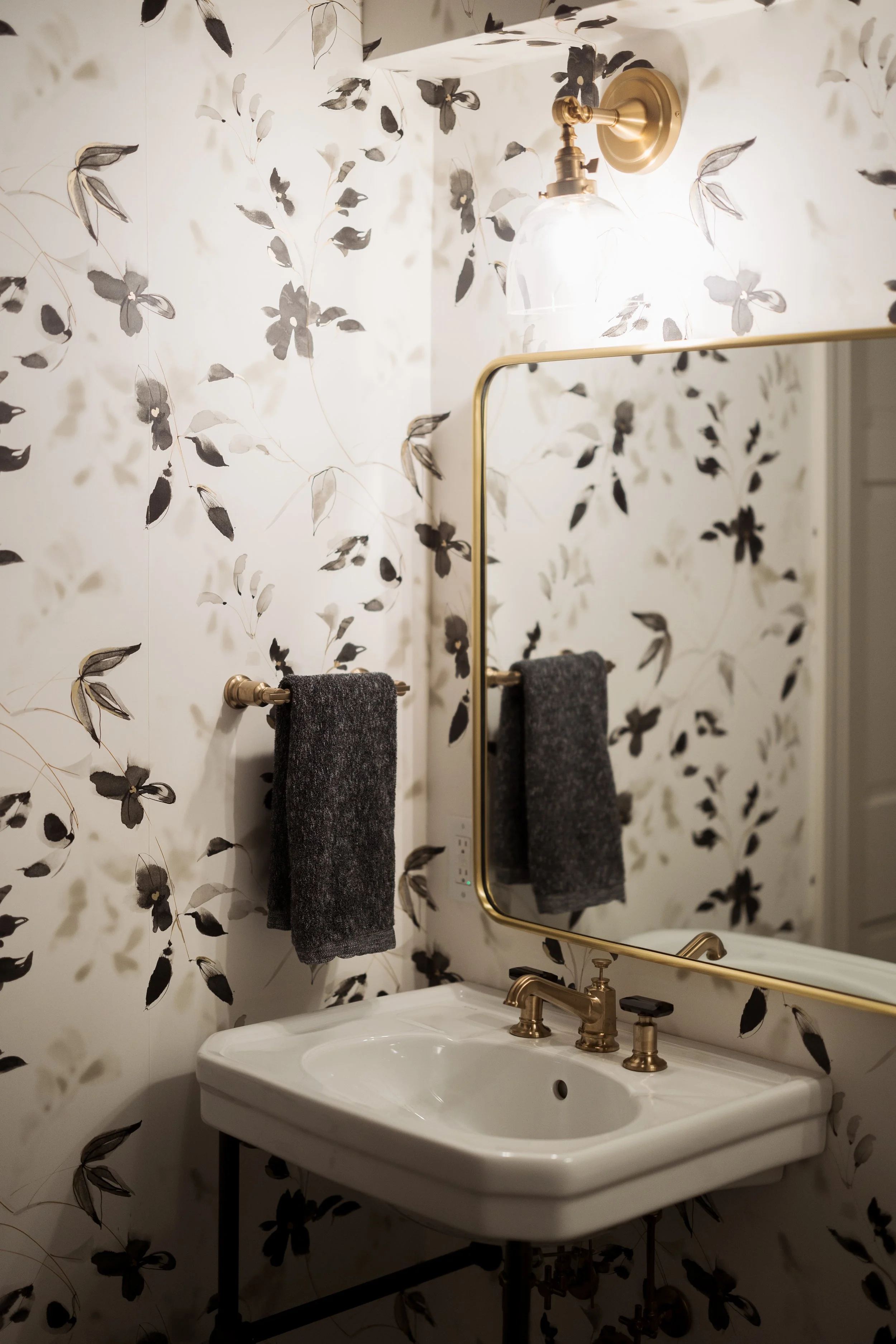 Bathroom sink with gold faucet and floral wallpaper, gold-framed mirror, and wall-mounted light fixture.