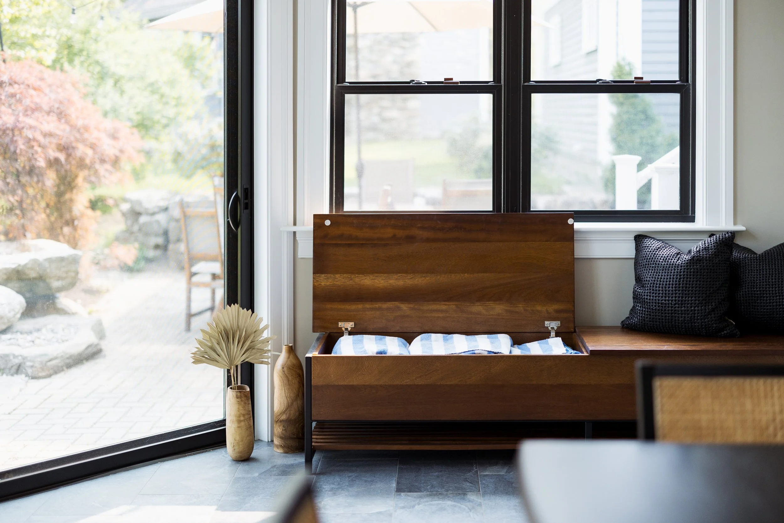 A modern living room with a wooden storage bench, open to reveal blue and white striped blankets. Next to it are decorative vases with dried plants. The space is bright with large windows and a view of a patio with outdoor furniture.