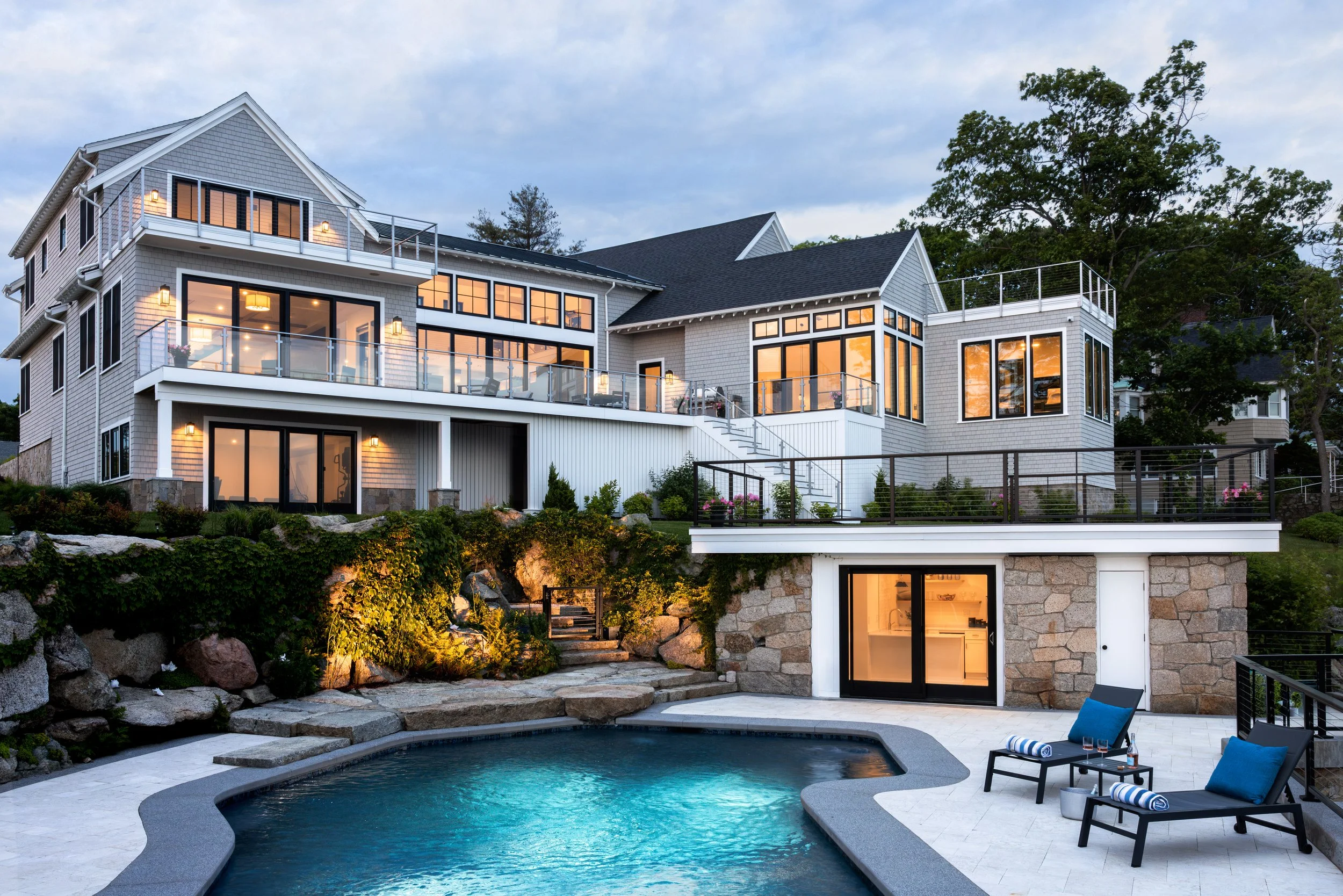 Modern luxury house with large windows and multiple levels, featuring a lit swimming pool, stone retaining walls, outdoor lounge chairs with blue cushions, and a glass railing.