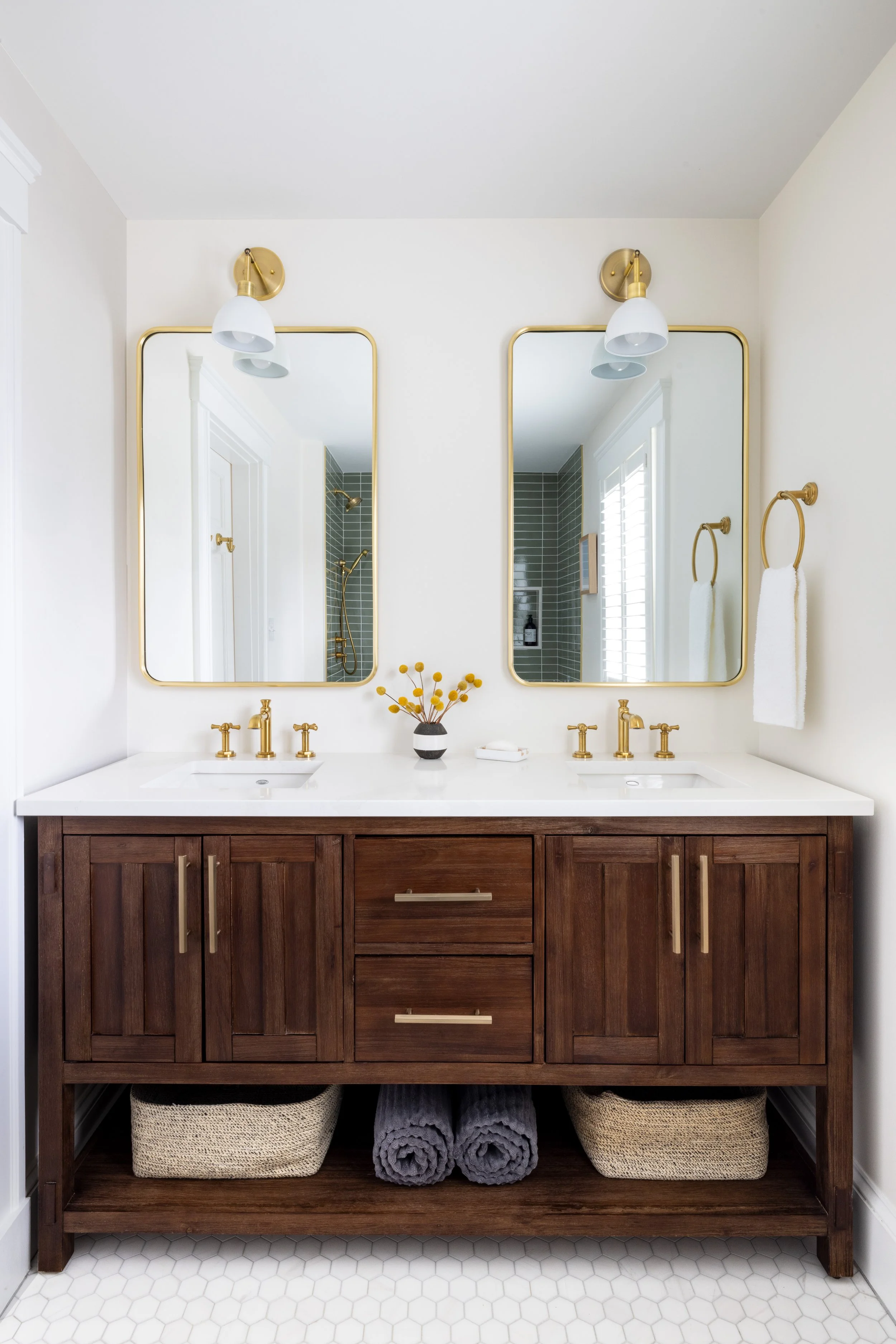 Modern bathroom with double vanity, two mirrors, gold fixtures, wooden cabinet, folded towels, and decorative accents.