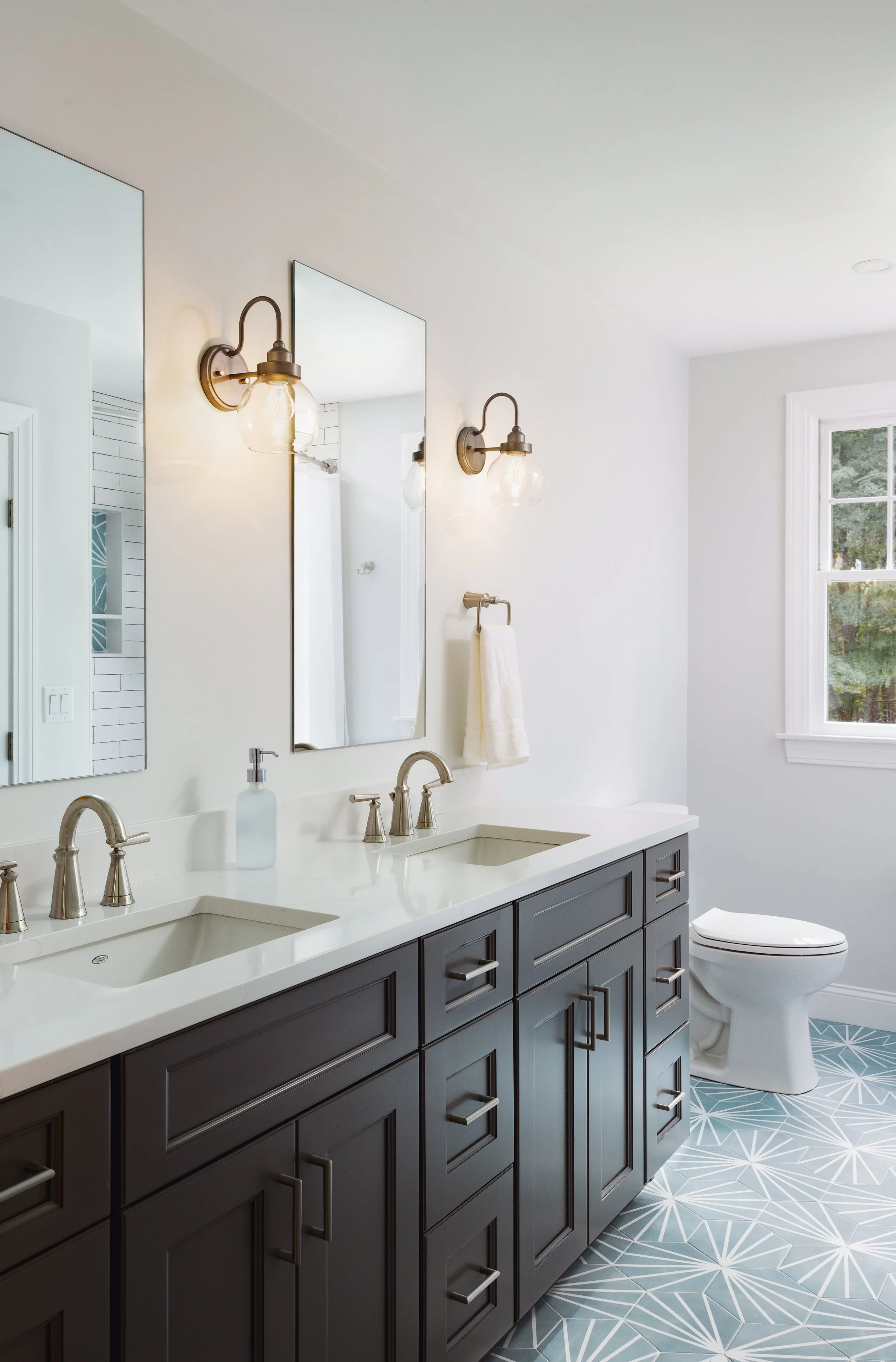 Modern bathroom with double sink vanity, dark cabinetry, two mirrors, wall-mounted lights, and patterned tile floor.
