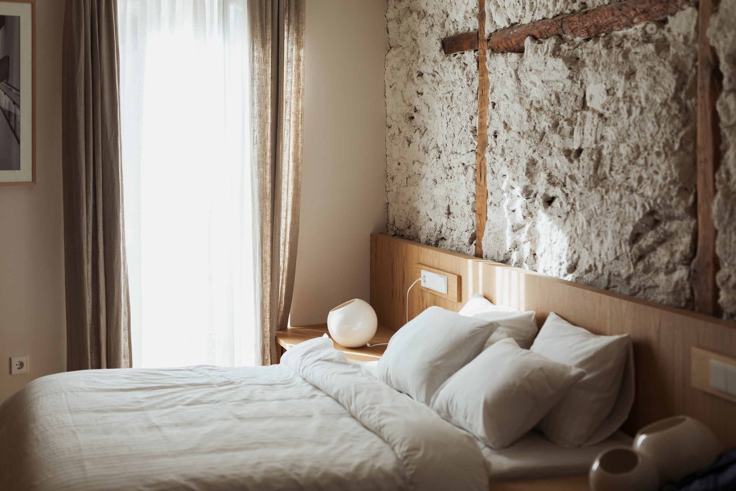 Cozy bedroom with a double bed, white bedding, rustic stone wall, large window, beige curtains, wooden headboard, and modern lamp.