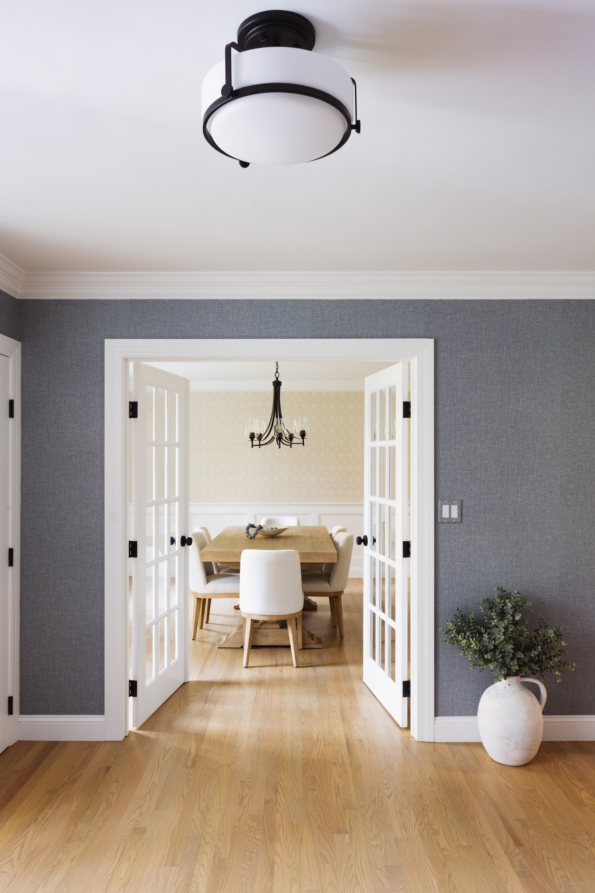 Modern dining room with wooden table, upholstered chairs, chandelier, and potted plant, viewed through French doors.