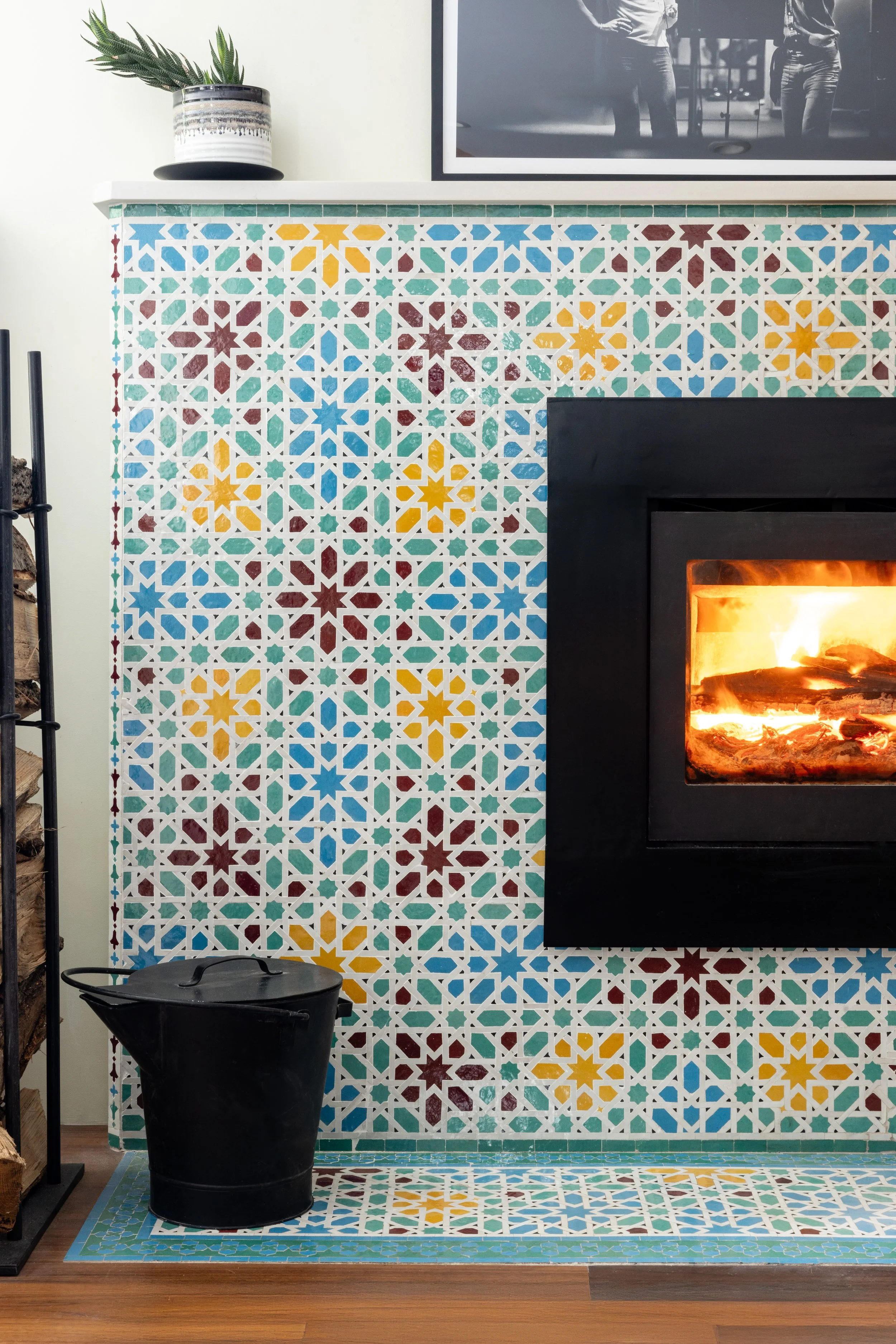 Colorful patterned tiles around a black fireplace with a fire burning inside; a black bucket sits on the wooden floor next to the fireplace, and a plant is on the mantel above. A black and white photo is hung above the mantel.