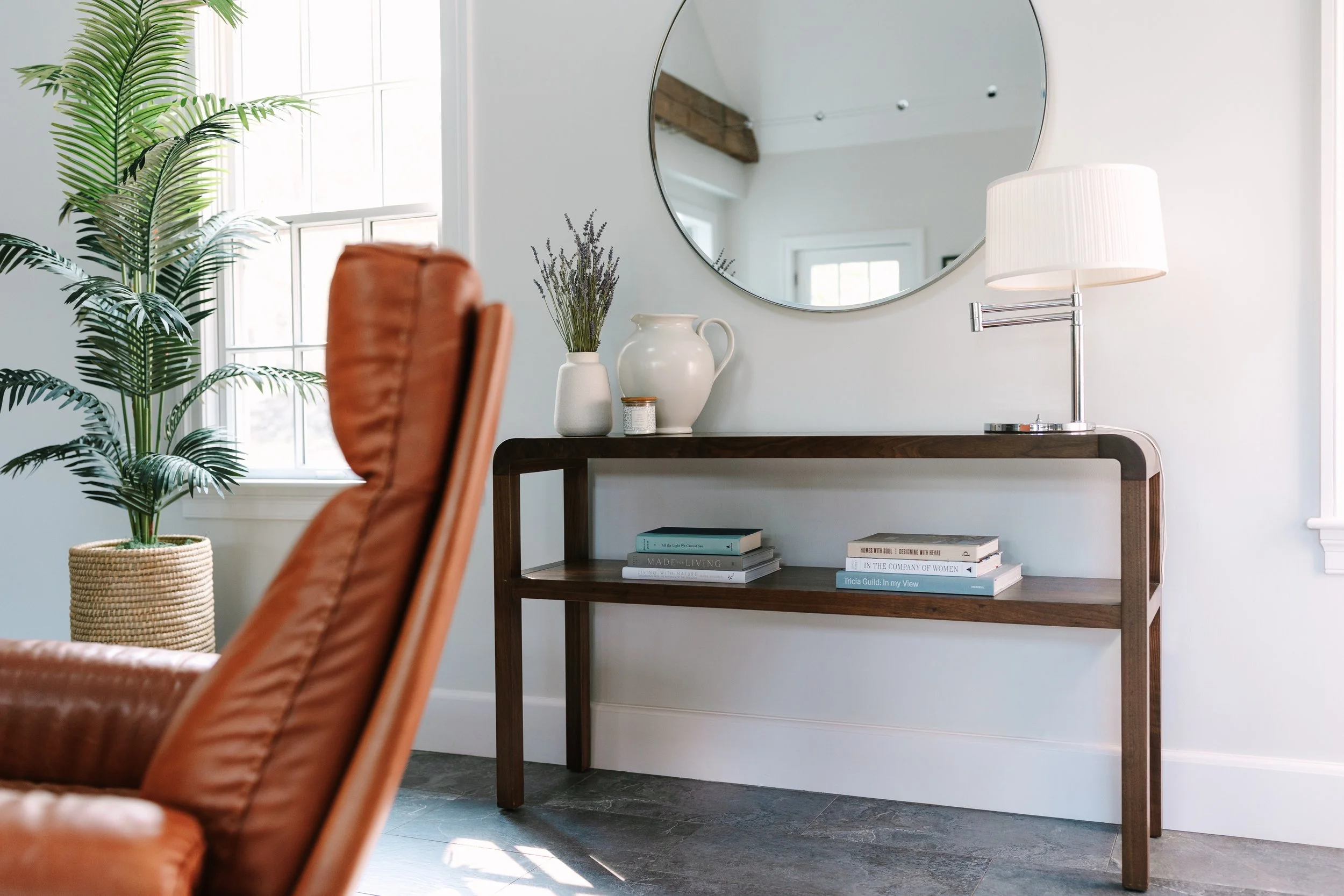 Modern living room with a wooden console table, a round mirror, and a leather chair. Decor includes a potted plant, decorative vase, lamp, and stacked books.