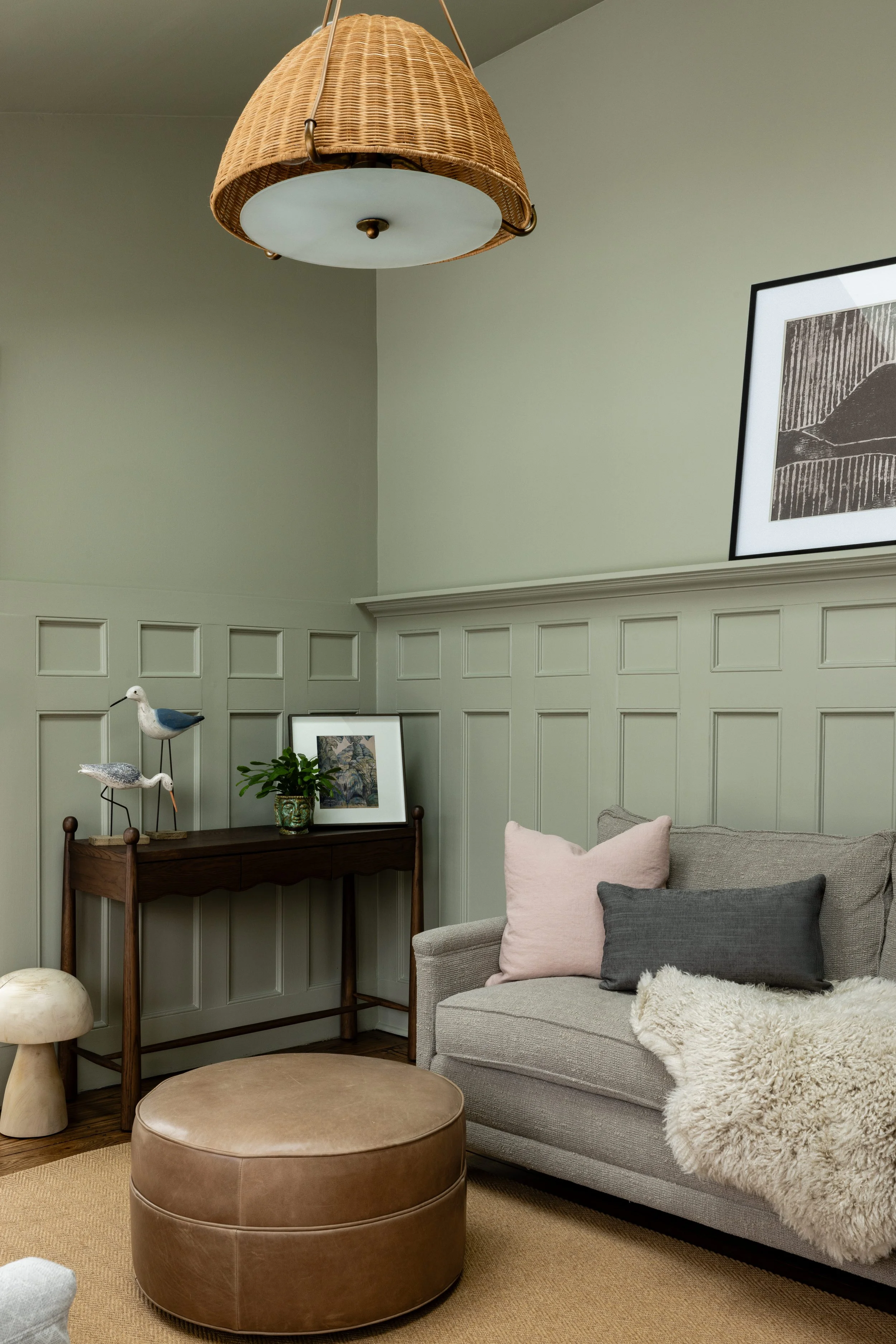 Cozy living room with a beige sectional sofa, topped with pink and gray cushions and a fur throw, situated beside a wooden console table. The table holds decorative items including bird sculptures and a framed picture, against sage green wainscoting.