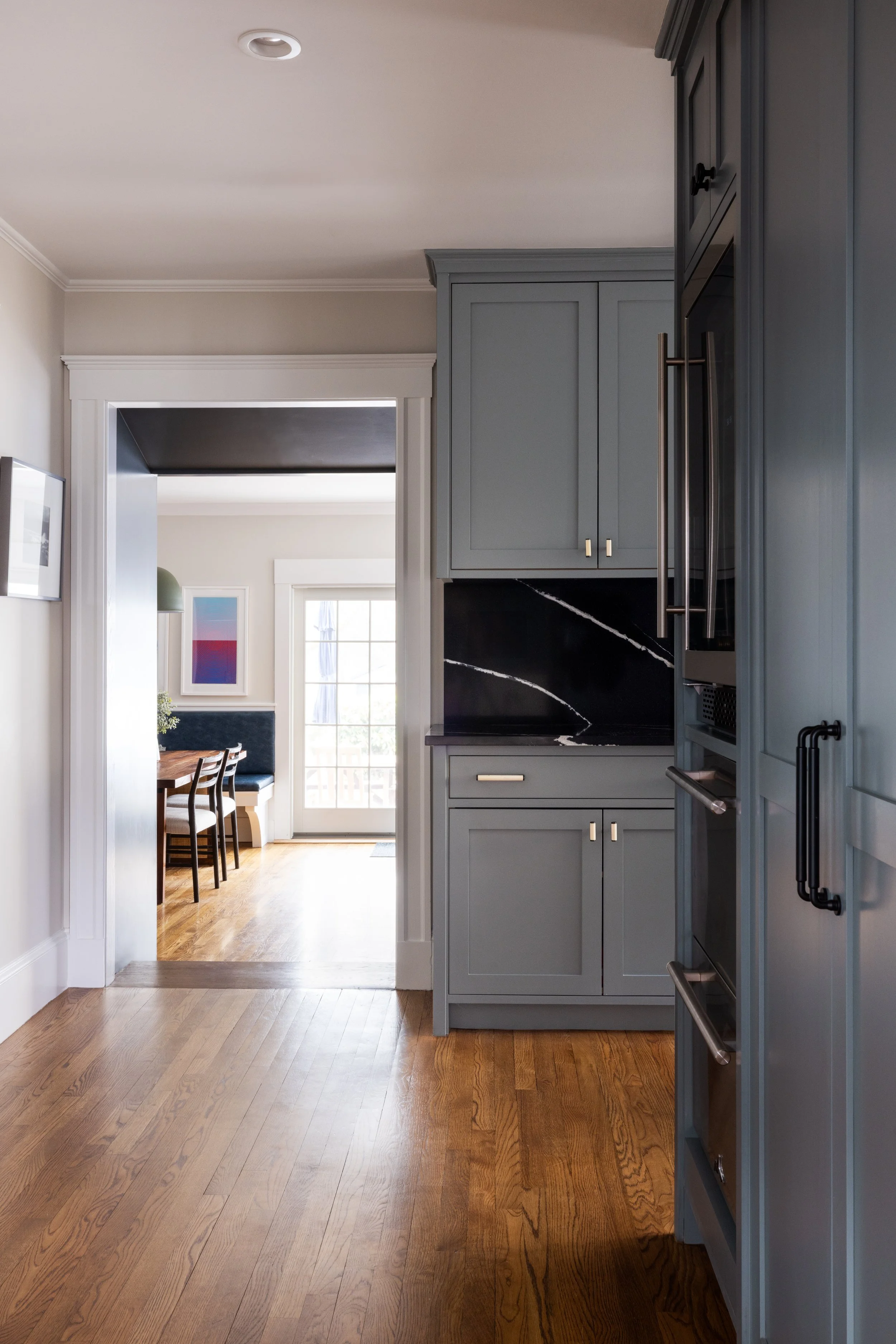 View of a modern kitchen with blue-gray cabinets, black marble backsplash, and stainless steel appliances, leading to a dining area with a wooden table and chairs, a blue cushioned bench, artwork on the wall, and a glass door leading outside.