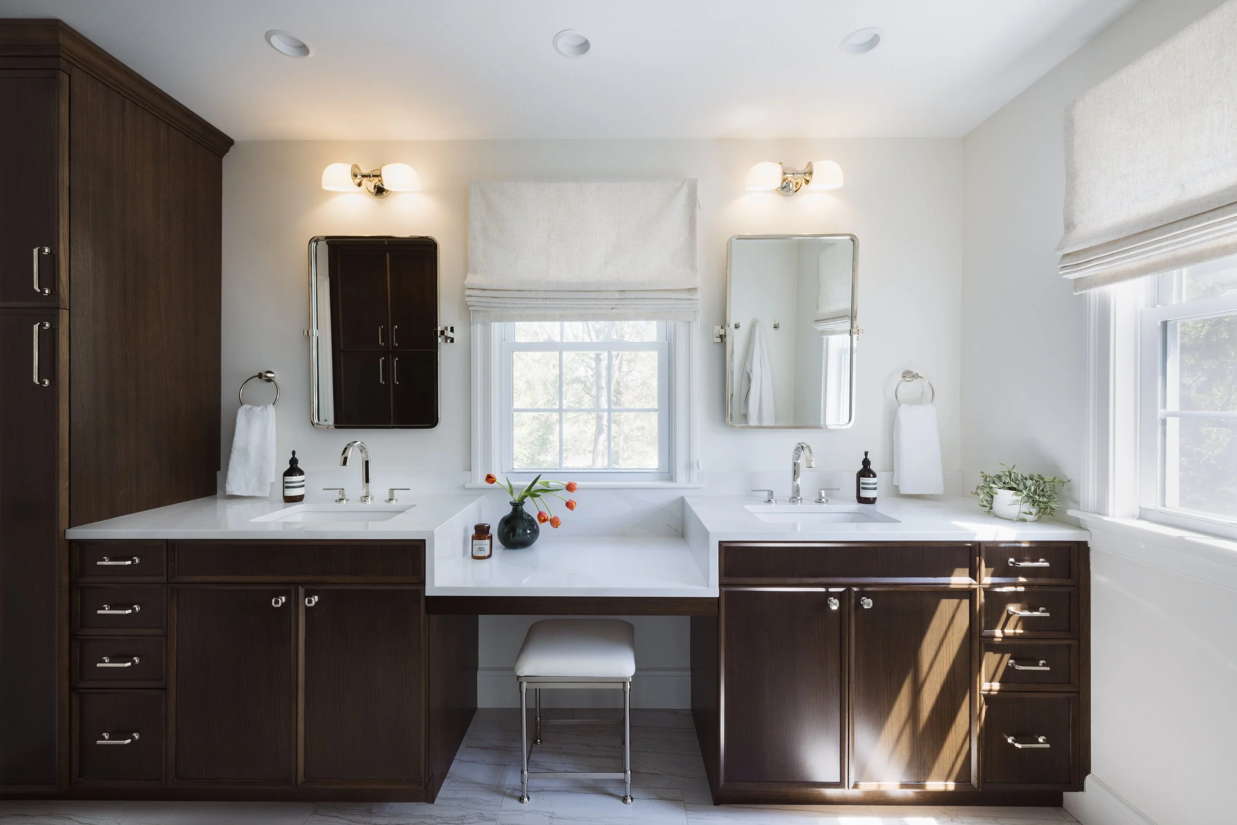 Modern bathroom with double sink vanity, dark wood cabinets, white countertops, and wall mirrors. Features beige window shades, a small stool, a vase with orange flowers, soap dispensers, and a potted plant.