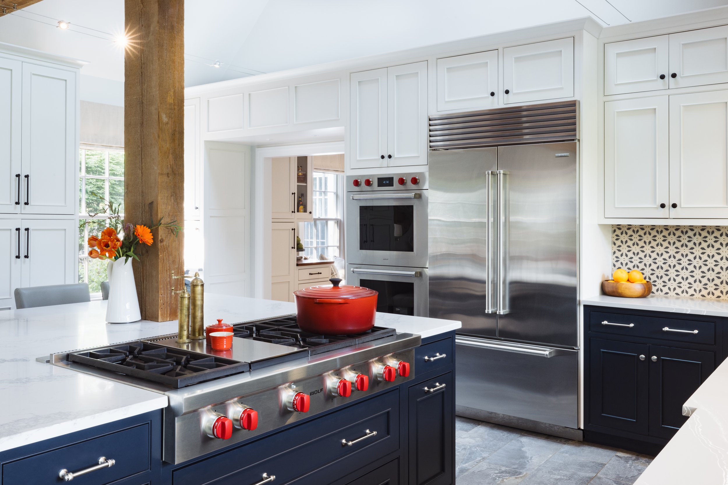 Modern kitchen with white cabinets, stainless steel fridge, and a center island featuring a stove with red knobs. A red pot is on the stove, and there's a white vase with orange flowers on the counter. The backsplash has a geometric pattern, and a wo