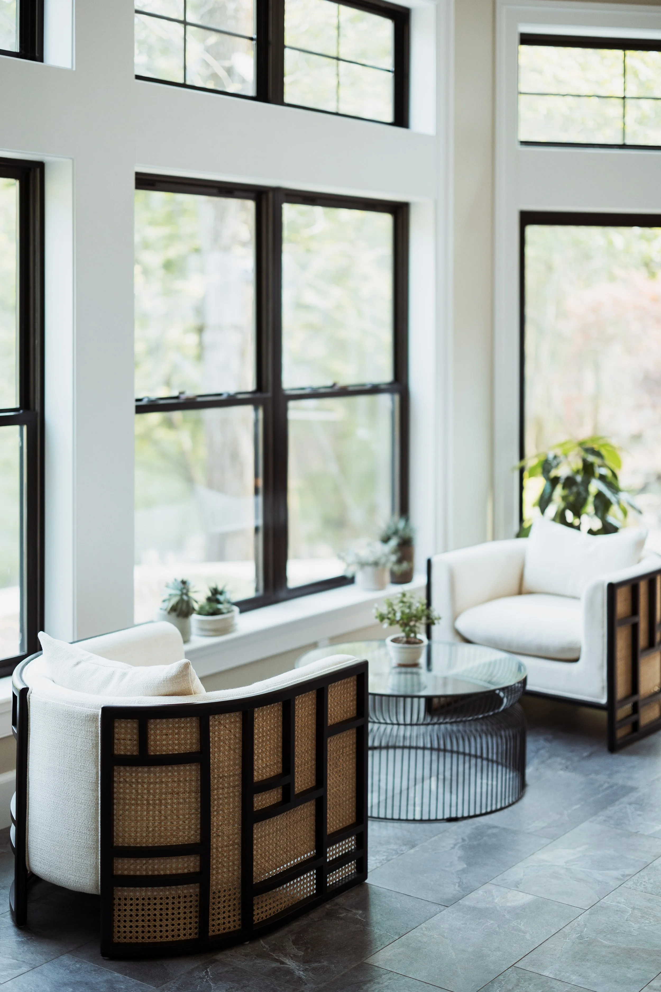 A modern living room with two beige armchairs and a round glass coffee table. The room has large windows with black frames, allowing natural light to illuminate the space. Small potted plants are placed on the windowsill and table, adding greenery to