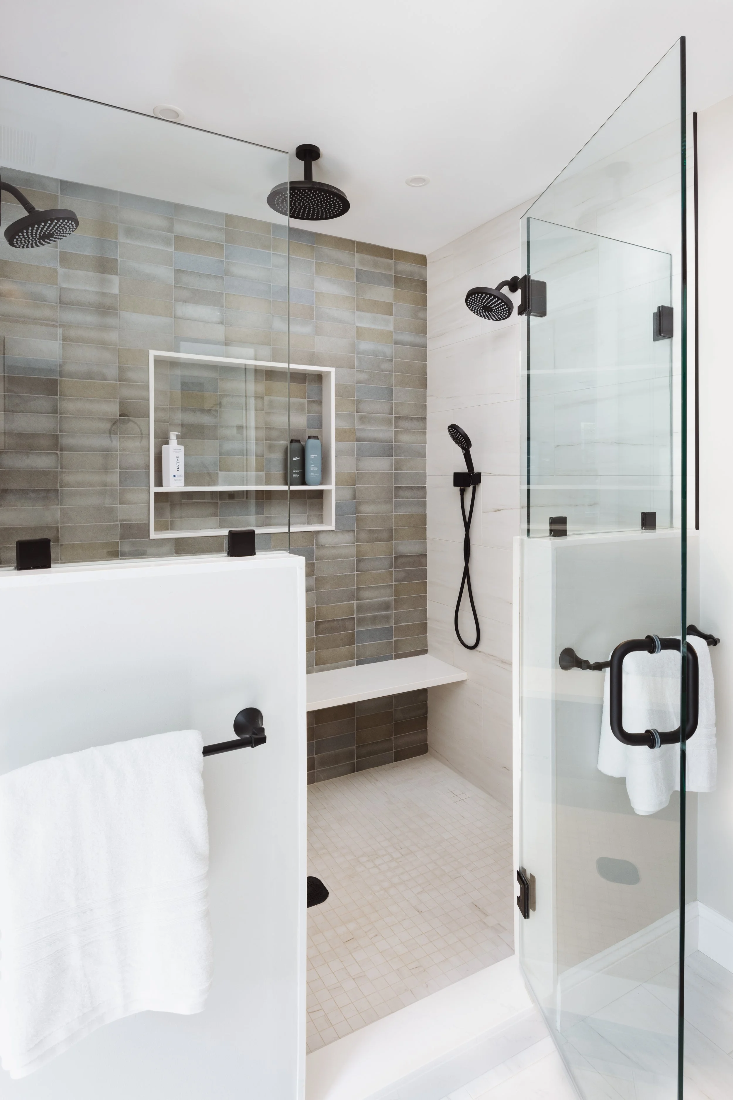 Modern bathroom with glass-enclosed shower, dual shower heads, beige tiles, built-in shelf with toiletries, and a white towel on a black rack.