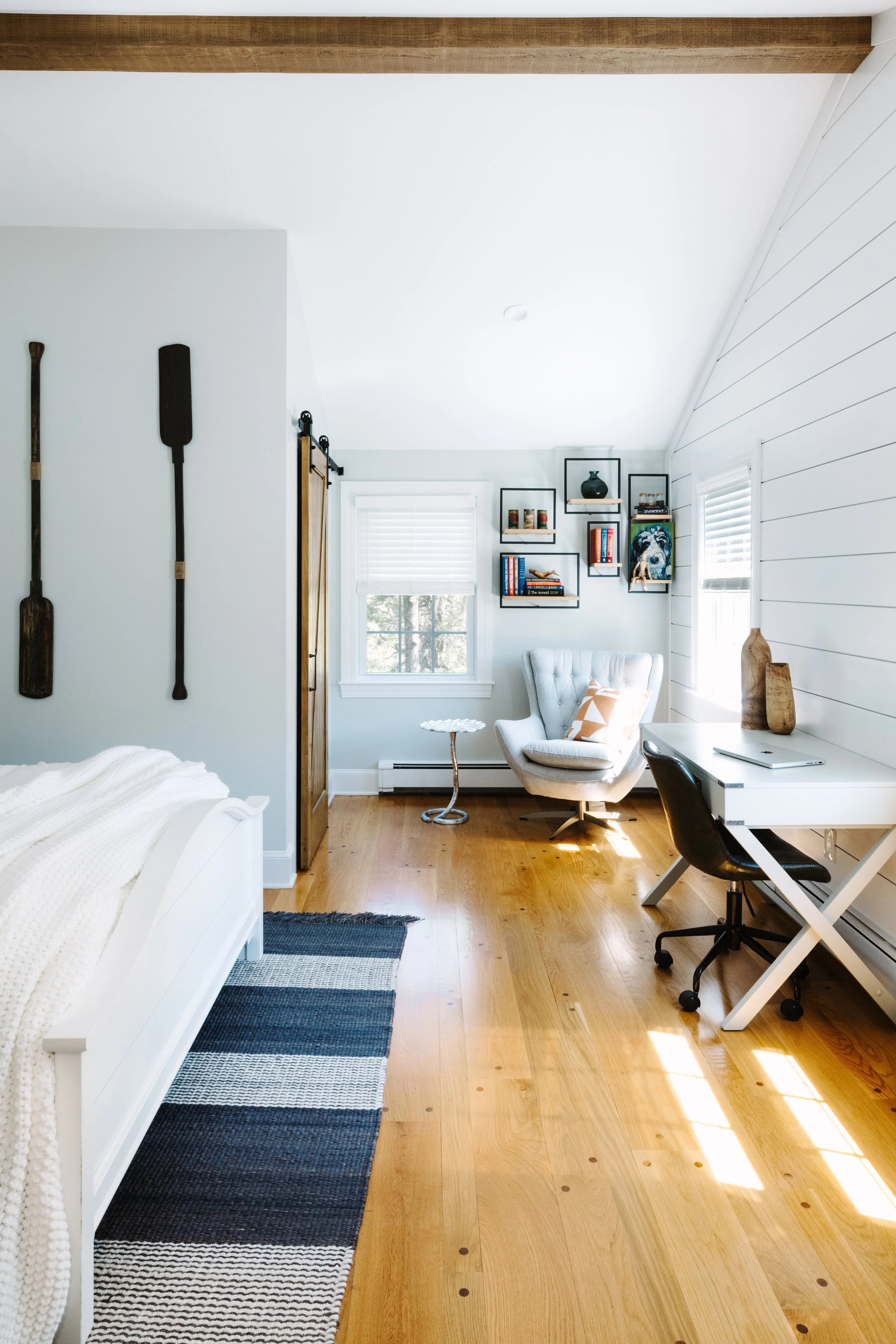 Cozy bedroom with wooden flooring, featuring a bed with a white knit blanket, decorative oars on the wall, a sliding barn door, a gray upholstered chair with a geometric pillow, wall shelves with books and decor, a modern white desk, and a black offi