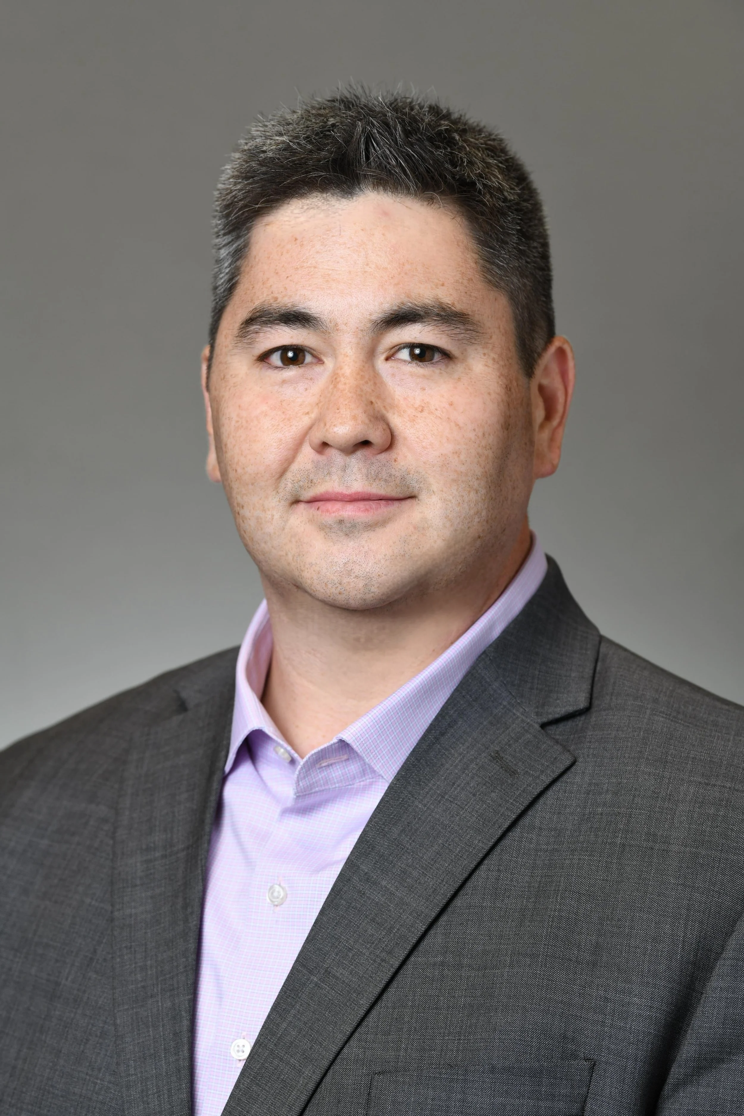 Professional headshot of a man with short dark hair, wearing a dark suit jacket and a light purple dress shirt against a gray background.