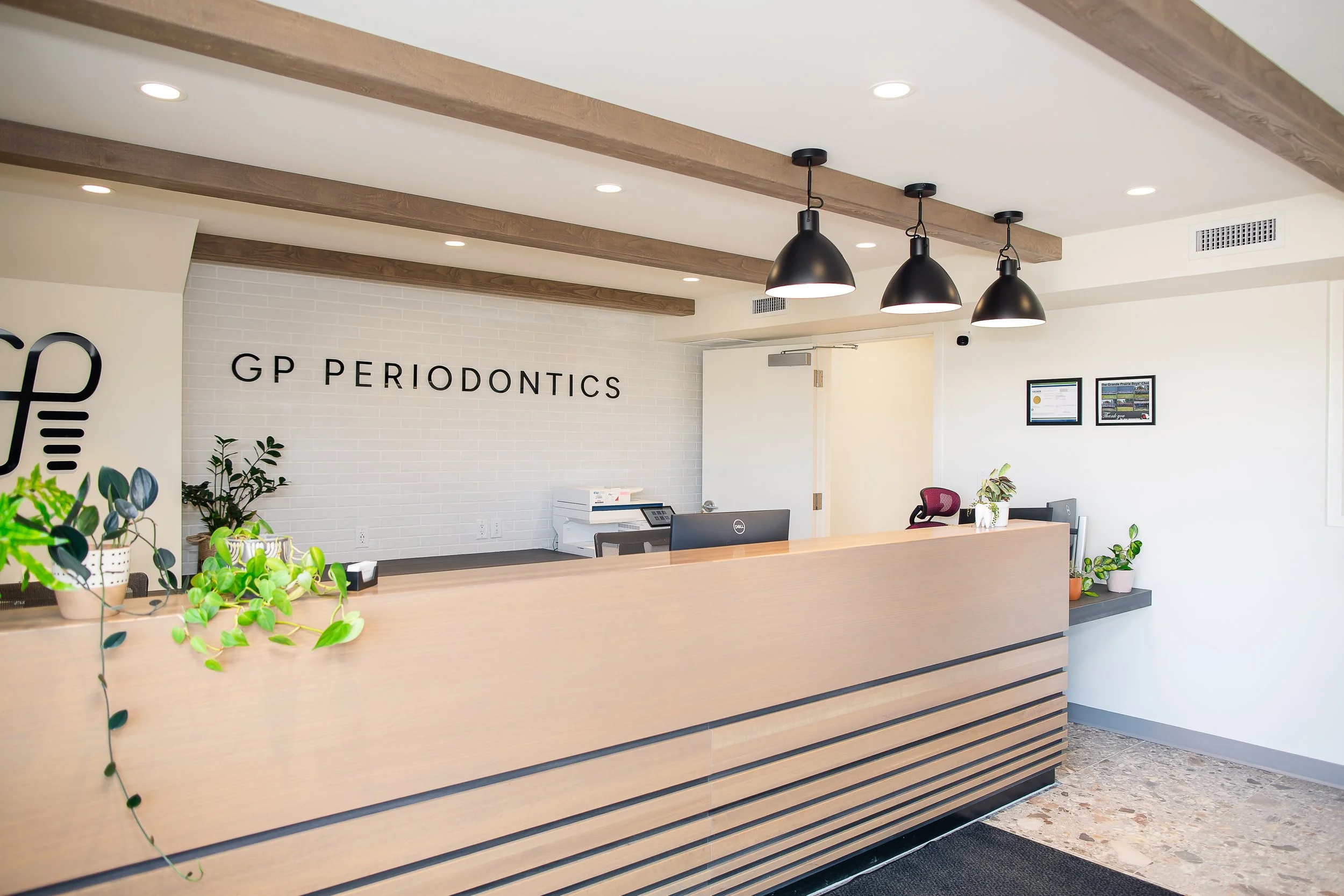 Reception desk in a modern dental clinic with plants and framed pictures, white brick wall with 'GP PERIODONTICS' sign, black hanging pendant lights, and wood ceiling beams.