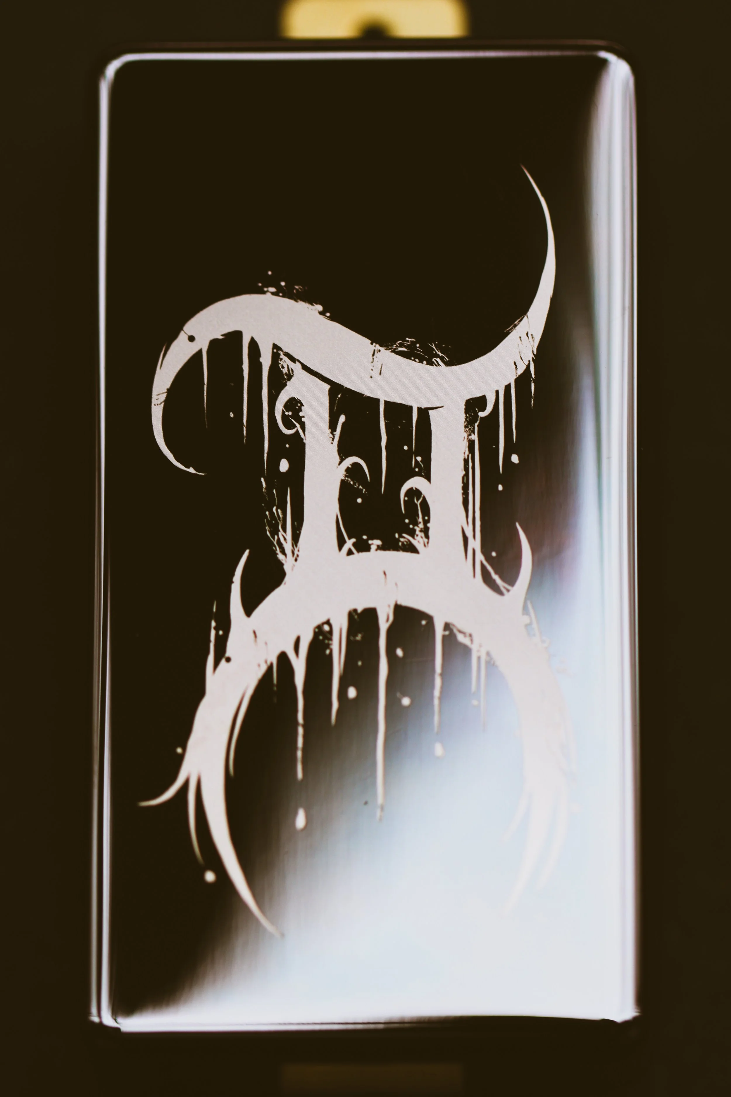 Macro photo of a single Arcana guitar pickup with a dripping black metal inspired Gemini symbol etched in white on a glossy black cover. Gold mounting hardware visible at the top.