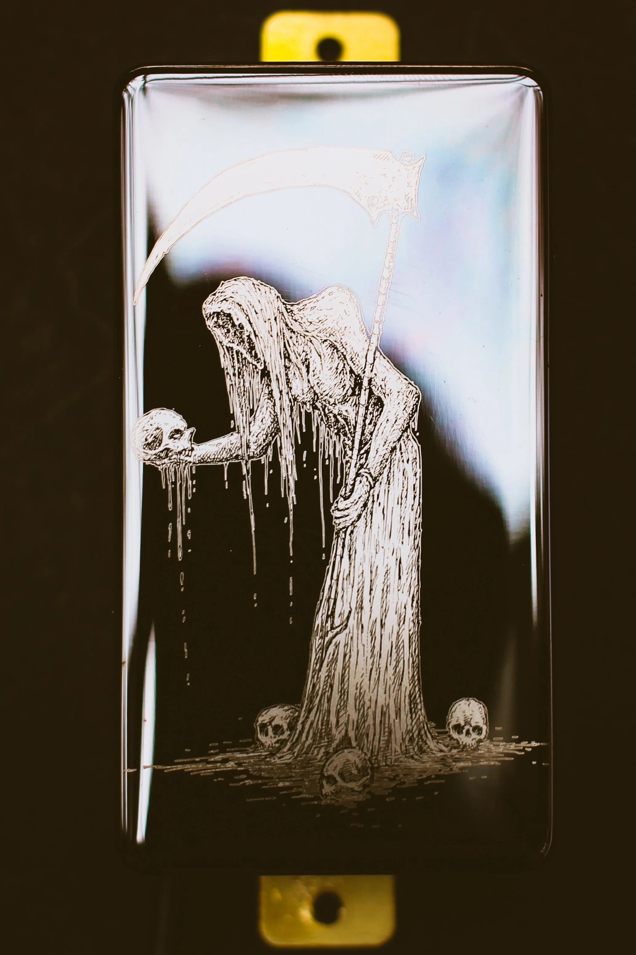 Close up of a custom Arcana guitar pickup featuring a detailed illustration of a robed reaper holding a skull, surrounded by small skulls at the base. White etching on a glossy black cover with gold mounting hardware.