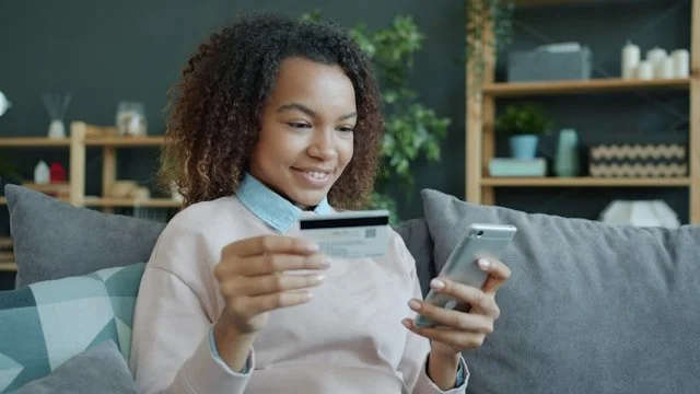Young woman looking at her phone and holding her card to do a conscious spending plan.