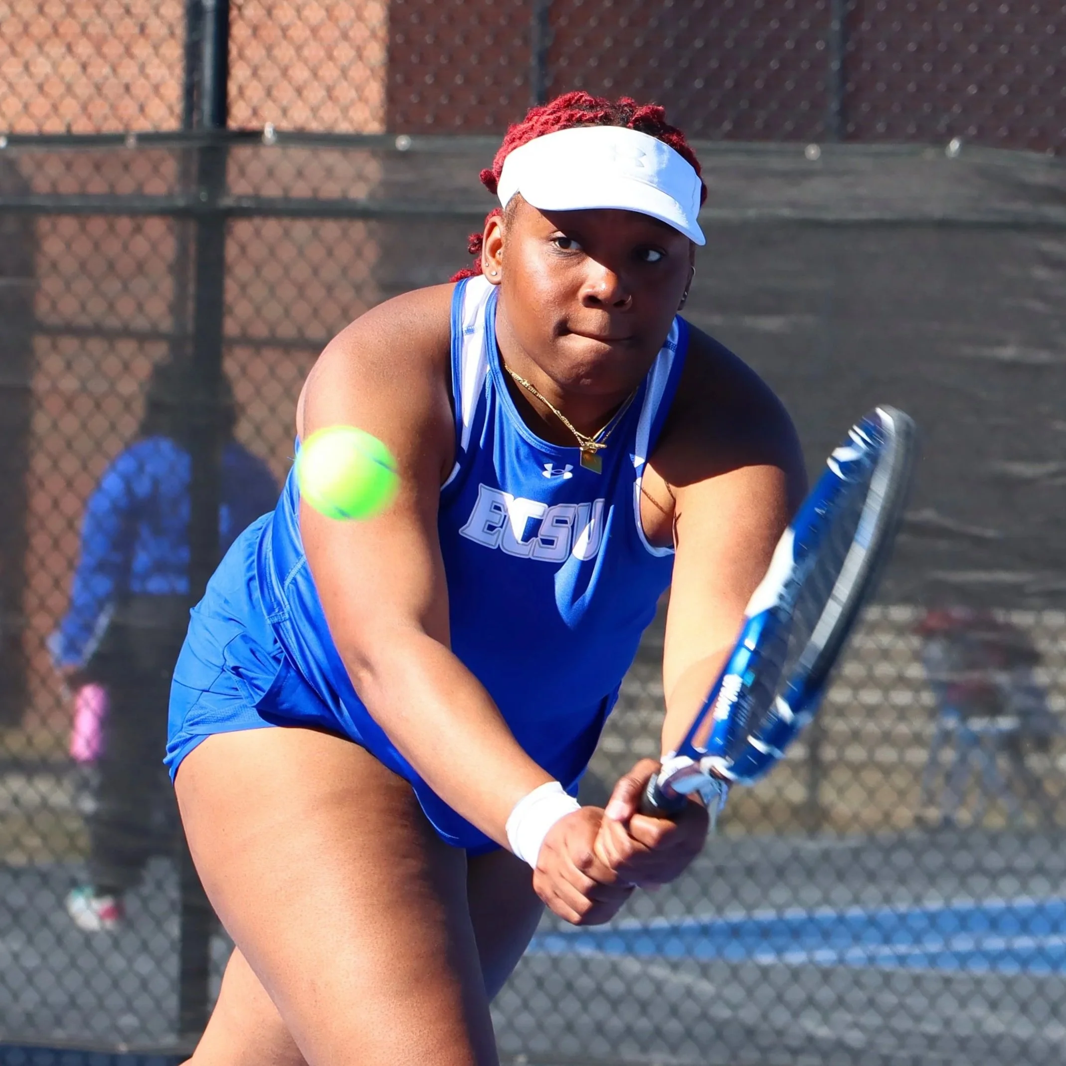 A young female tennis player in a blue Under Armour outfit and white visor preparing to hit a tennis ball on an outdoor court.