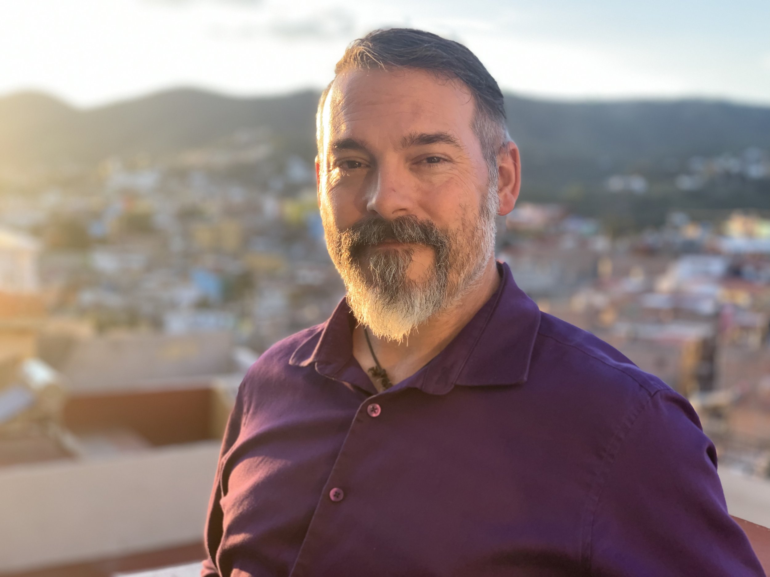 A man with a beard and mustache, wearing a purple shirt, poses outdoors during sunset with a blurred cityscape and hills in the background.