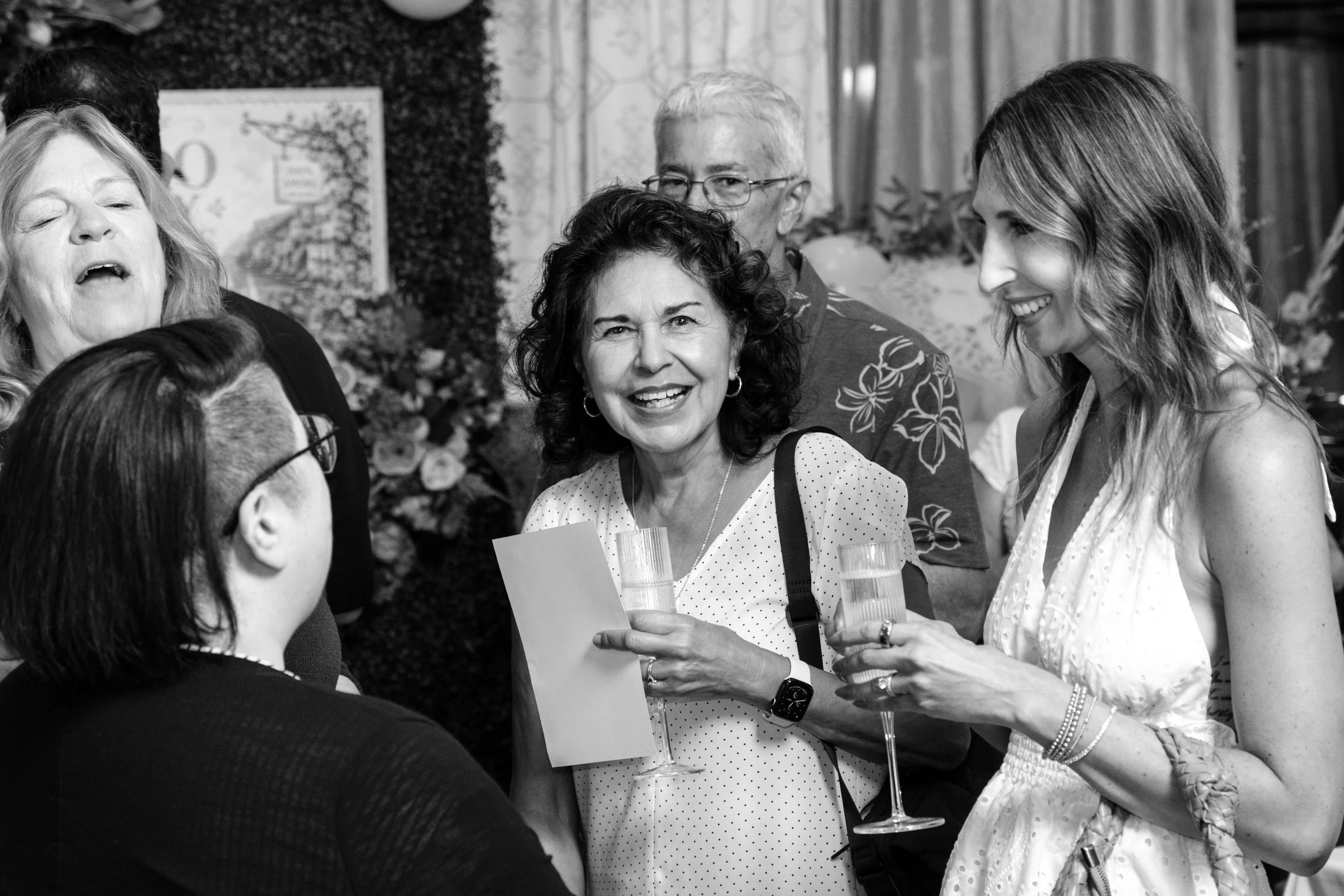 Group of people at a social gathering, smiling and holding drinks, engaging in conversation.