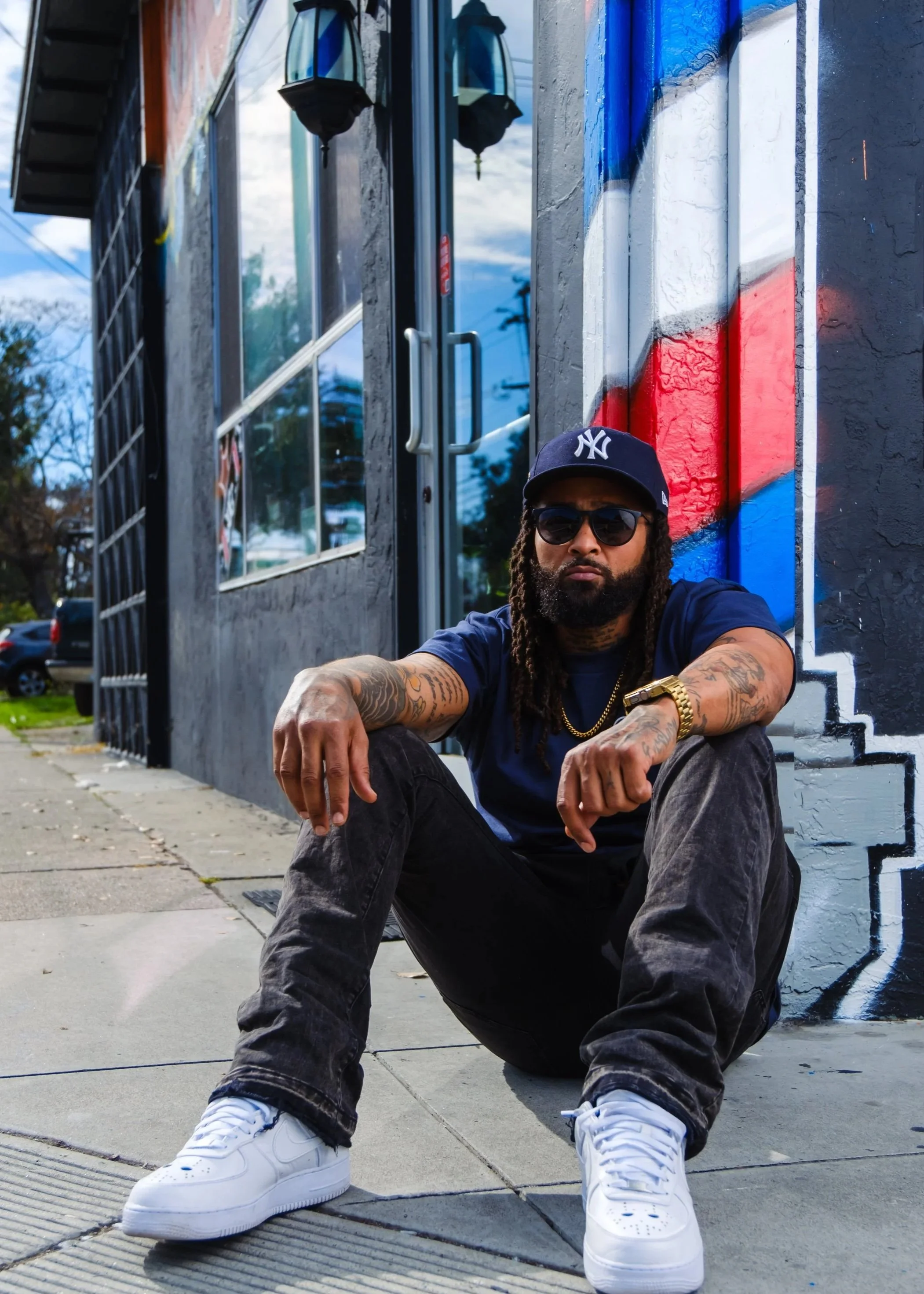 Man sitting on the sidewalk in front of a colorful mural, wearing sunglasses, a New York Yankees cap, gold chain, tattoos, and white sneakers.