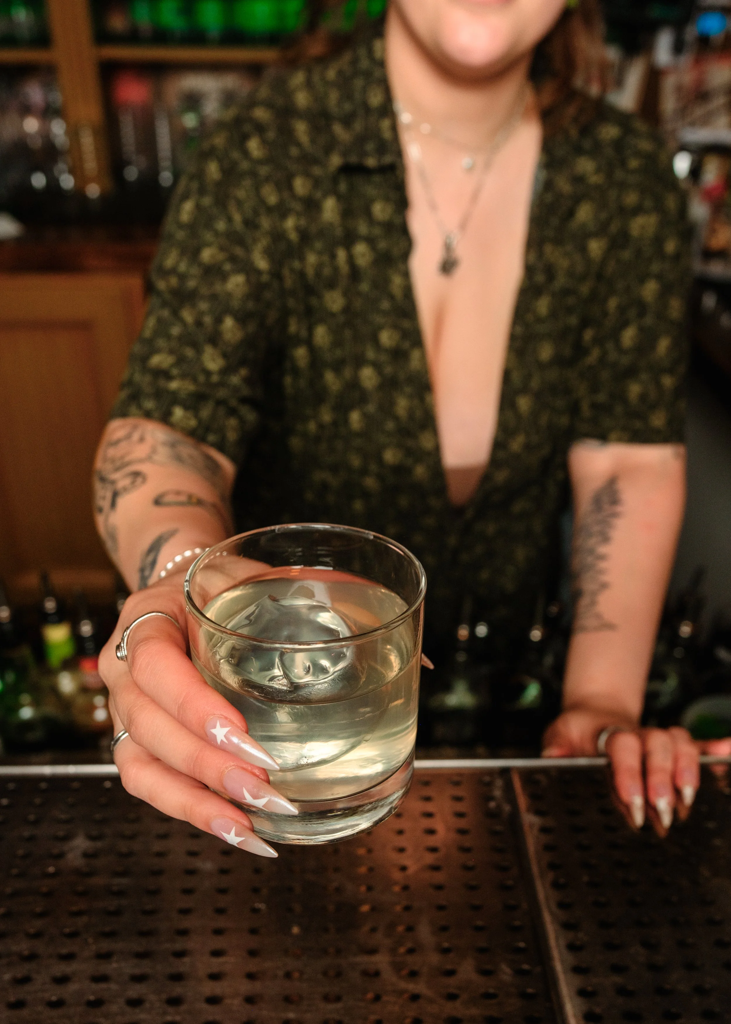 A woman with tattoos on her arms wears a patterned shirt and jewelry, holding a glass of clear liquid in front of her at a bar. The background shows shelves with bottles.